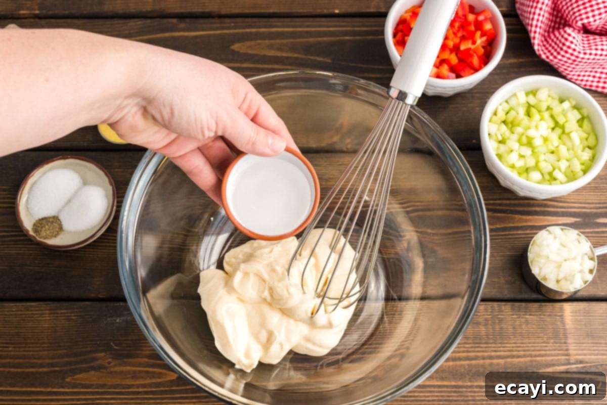 Nana's Homestyle Macaroni Salad 3 A measuring cup of white vinegar being poured into a large mixing bowl filled with mayonnaise, forming the base of the creamy dressing.