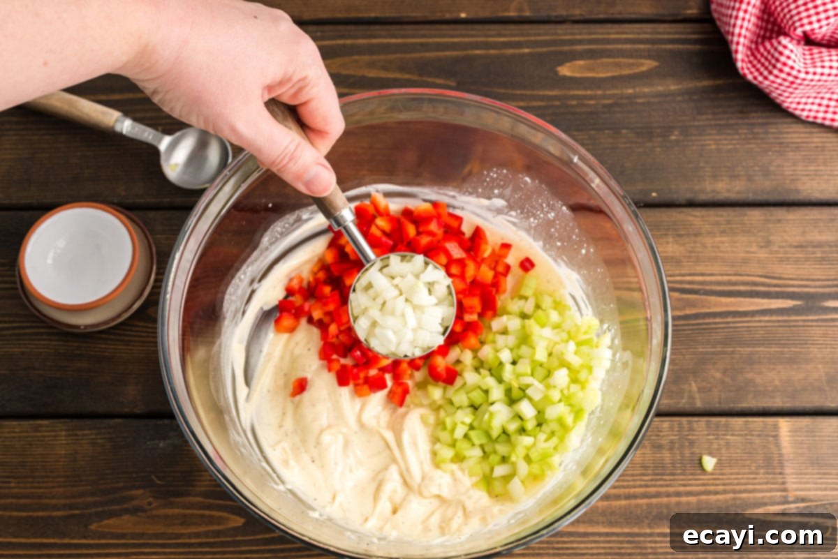 Nana's Homestyle Macaroni Salad 5 Finely diced onion being added to a large mixing bowl that already contains celery, bell peppers, and the prepared creamy dressing for the macaroni salad.