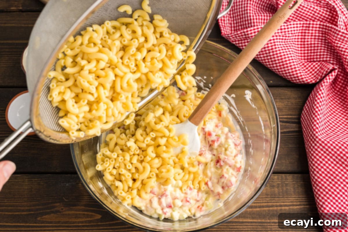 Nana's Homestyle Macaroni Salad 6 Cooked and cooled elbow macaroni being gently stirred into the large mixing bowl containing the creamy dressing and diced vegetables.