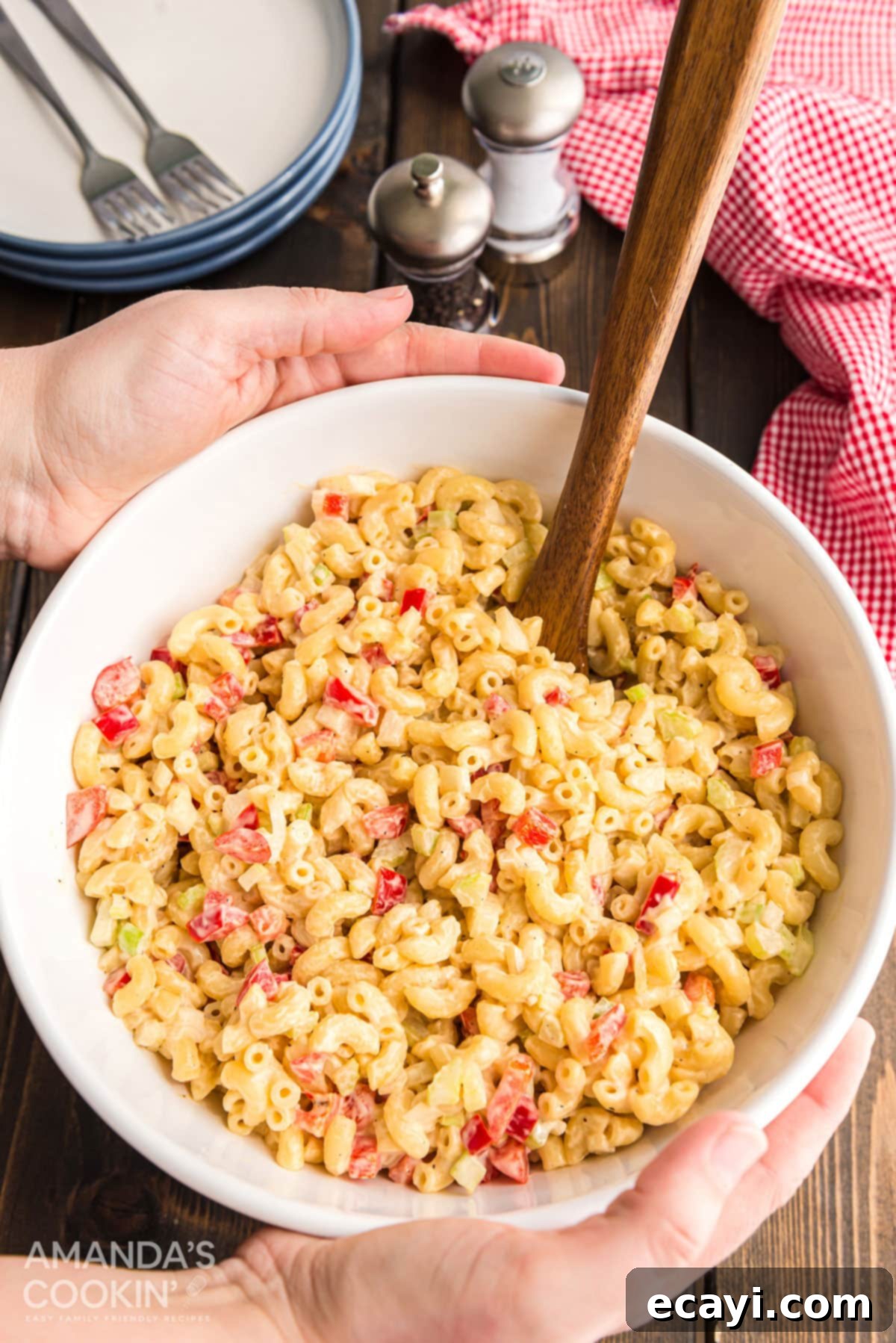 Nana's Macaroni Salad, perfectly chilled and ready to serve at any gathering. A pair of hands gently placing a large white bowl of creamy macaroni salad onto a beautifully set wooden table for serving, highlighting its portability and readiness.