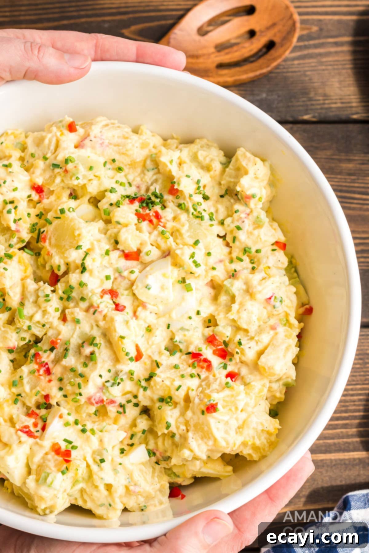 My mom's potato salad is a classic, creamy, and flavorful recipe that's the absolute hit of any party or potluck! Hands serving a large bowl of homemade potato salad with a wooden spoon, ready for a gathering.