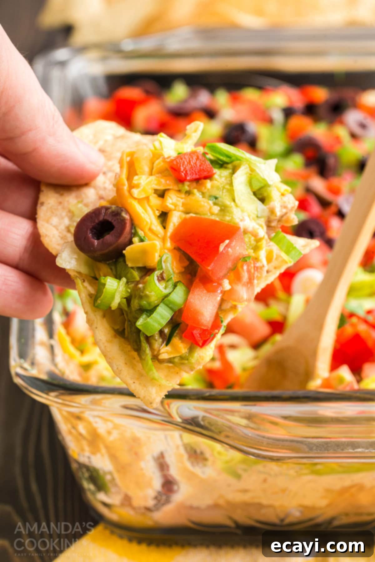 This ultimate 7-layer taco dip combines refried beans, creamy guacamole, seasoned sour cream, and a medley of fresh, crunchy toppings for the perfect party appetizer. Hand lifting a tortilla chip from a colorful, layered taco dip, showcasing fresh toppings