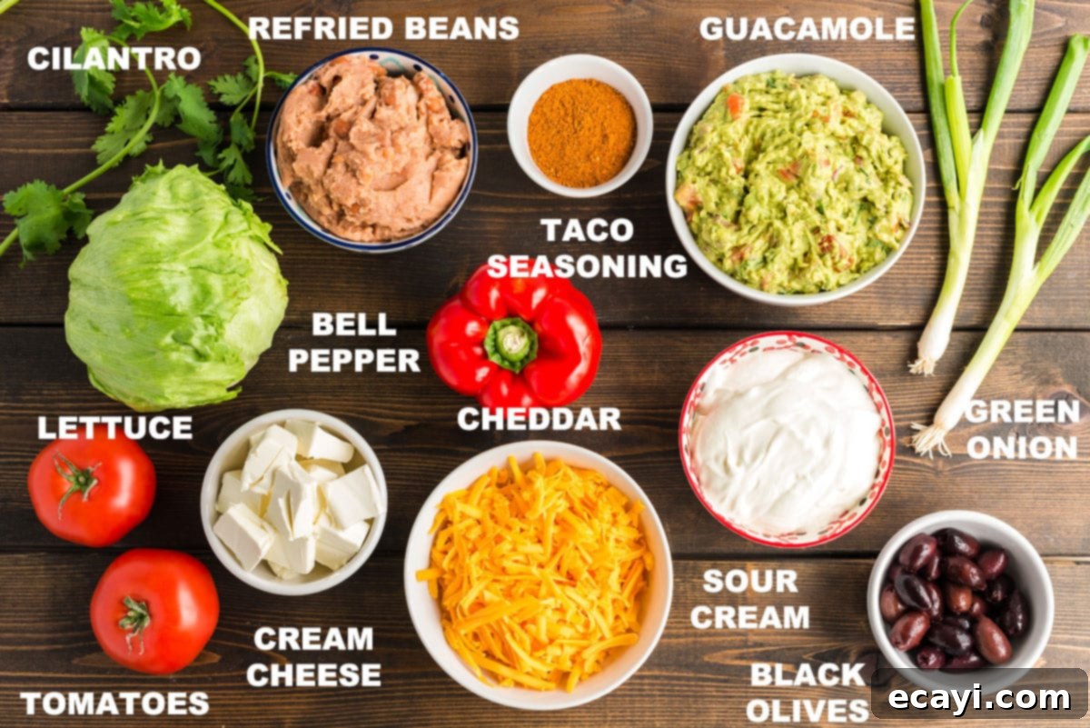 Ultimate Taco Dip 2 Labeled ingredients for a delicious 7-layer taco dip recipe, including refried beans, cream cheese, sour cream, guacamole, and fresh vegetables