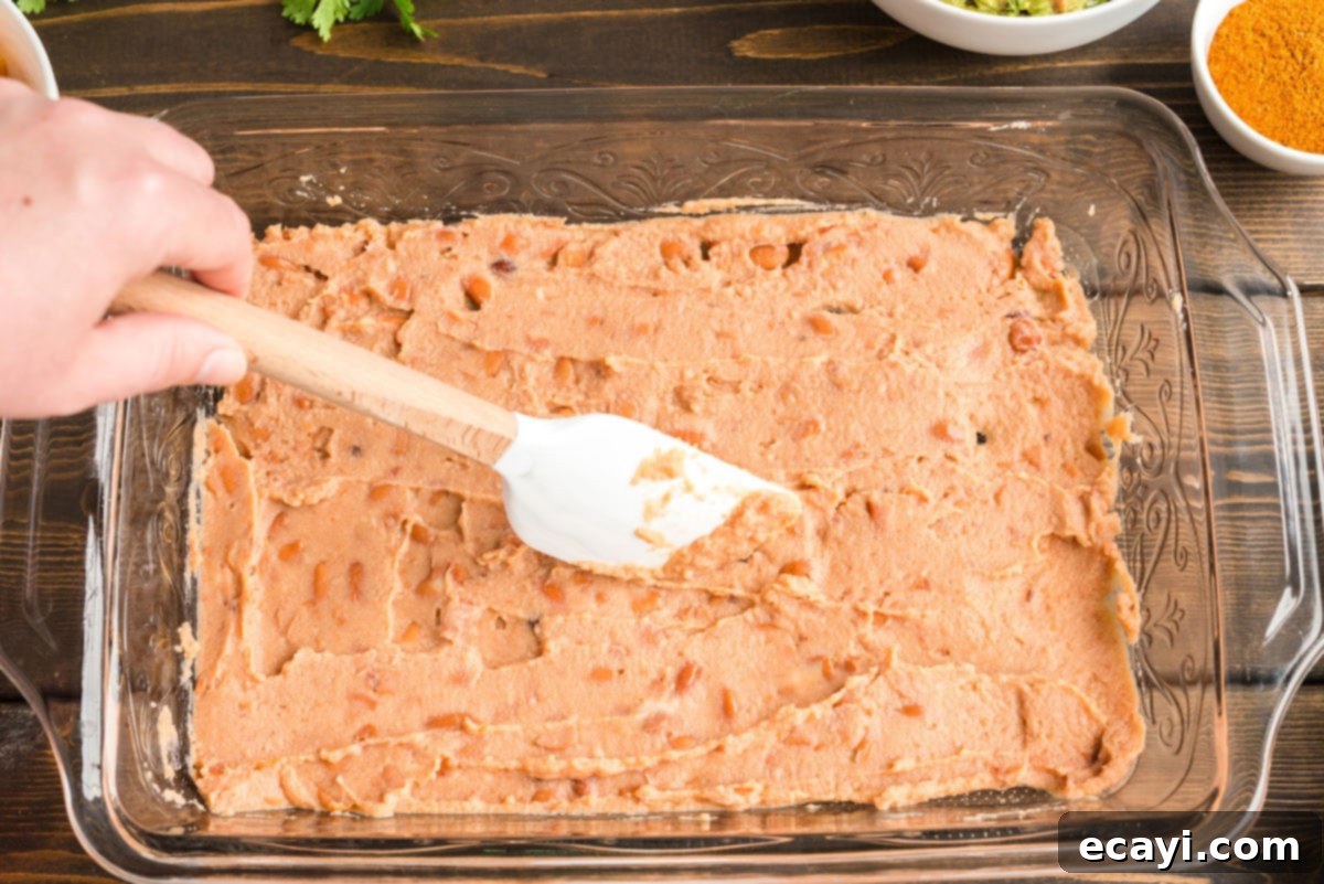 Ultimate Taco Dip 3 Spreading refried beans evenly into the bottom of a clear glass baking pan to form the first layer of taco dip