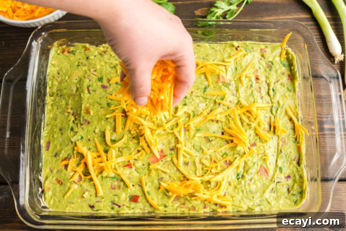 Ultimate Taco Dip 8 Sprinkling shredded cheddar cheese evenly over the guacamole layer in the taco dip, the first of the many fresh toppings