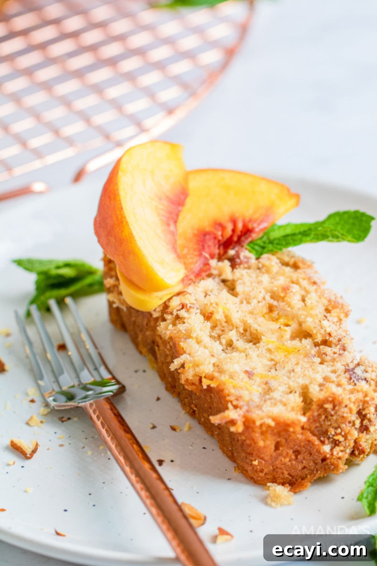 The delicious peach bread is studded with almonds and topped with an amazing cinnamon almond mixture you will love. A delectable slice of homemade peach quick bread, studded with almonds and topped with a cinnamon almond crumble, resting invitingly on a white plate next to a fork, ready to be enjoyed.