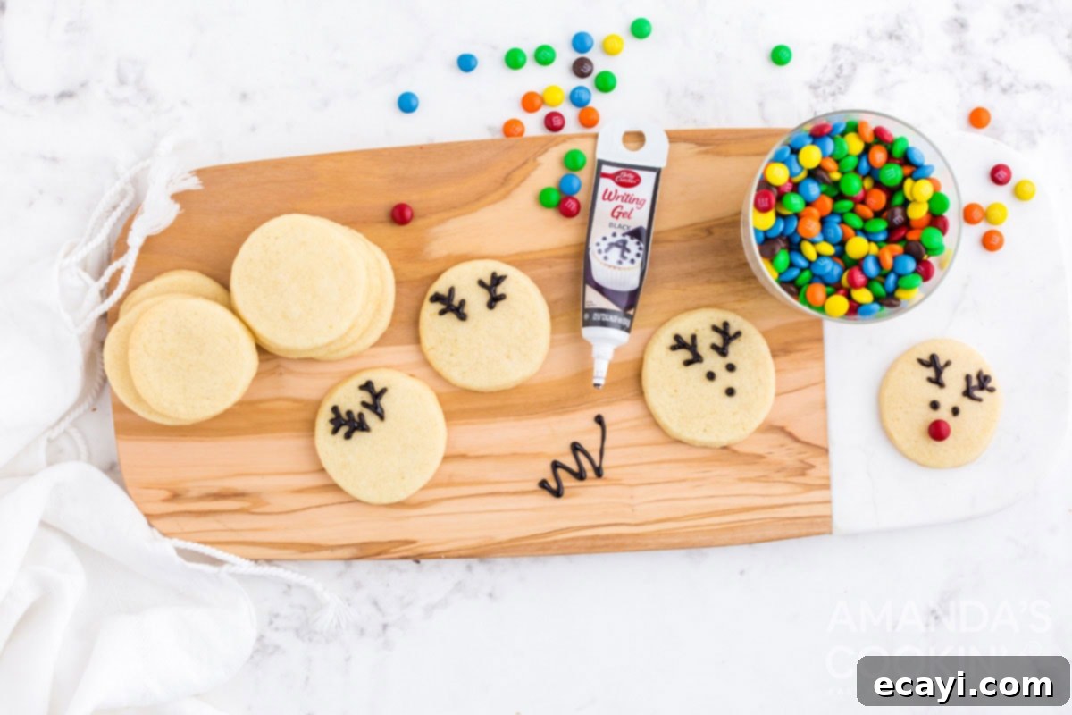 Sweet & Simple Decorated Cookies 23 Close-up of a hand using black decorator gel to draw antlers and eyes on a plain sugar cookie, next to mini M&Ms.
