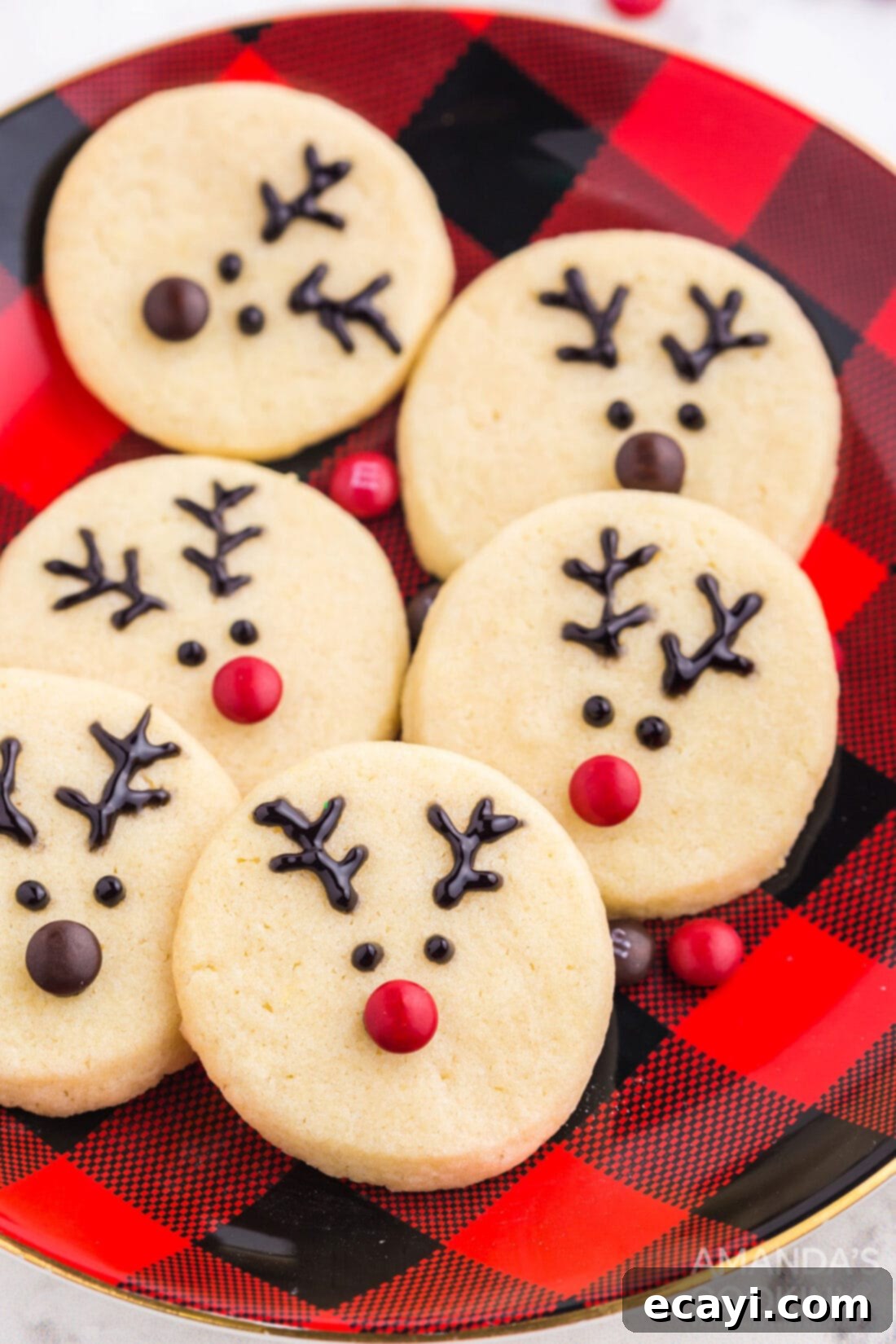 Sweet & Simple Decorated Cookies 4 Reindeer-themed sugar cookies, featuring brown and red mini M&M noses and drawn antlers, arranged on a red and black plaid plate.