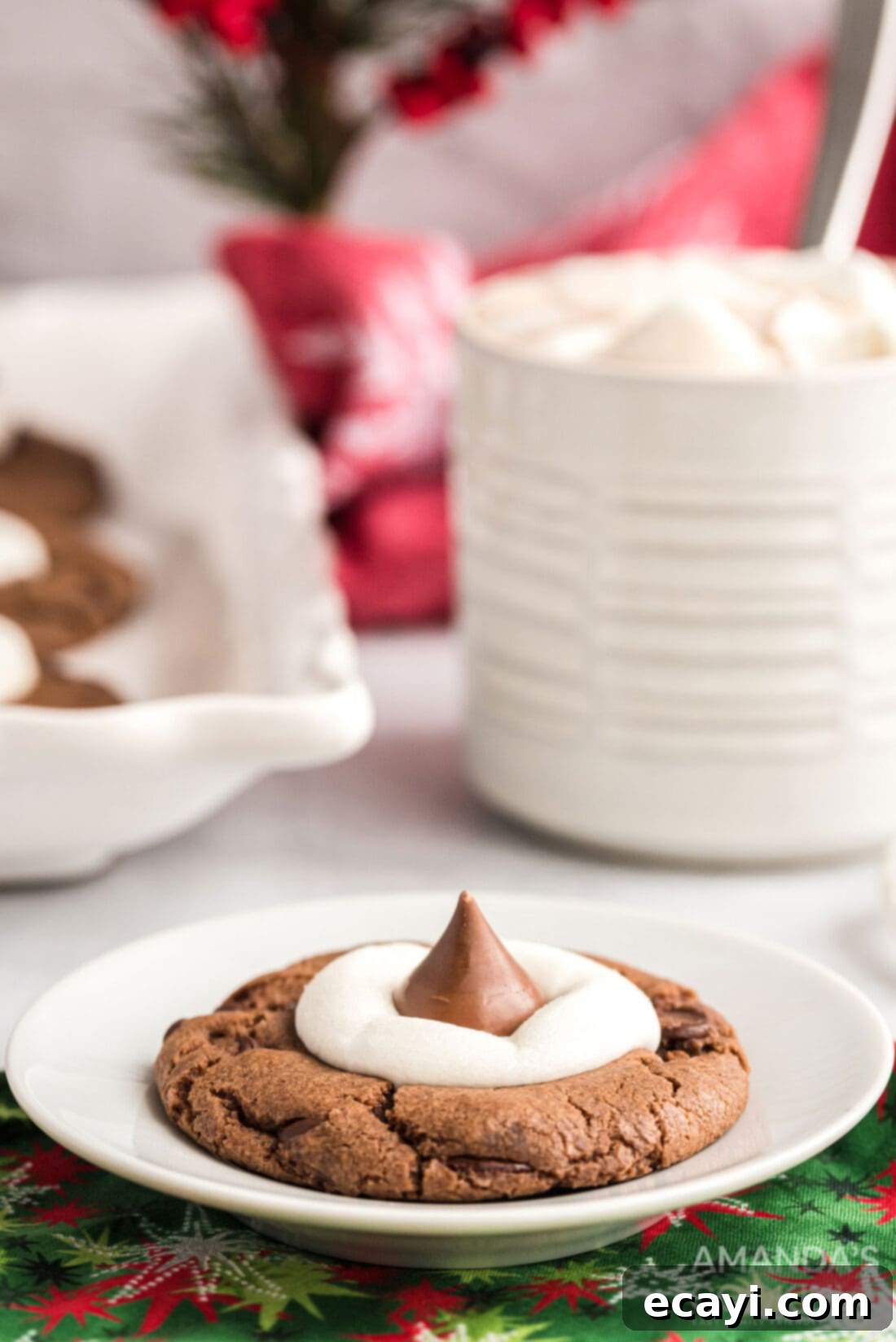 Cozy Cocoa Cookies 16 A single hot cocoa cookie with a melted marshmallow and chocolate shavings on a white plate, with a festive mug of hot chocolate blurred in the background.