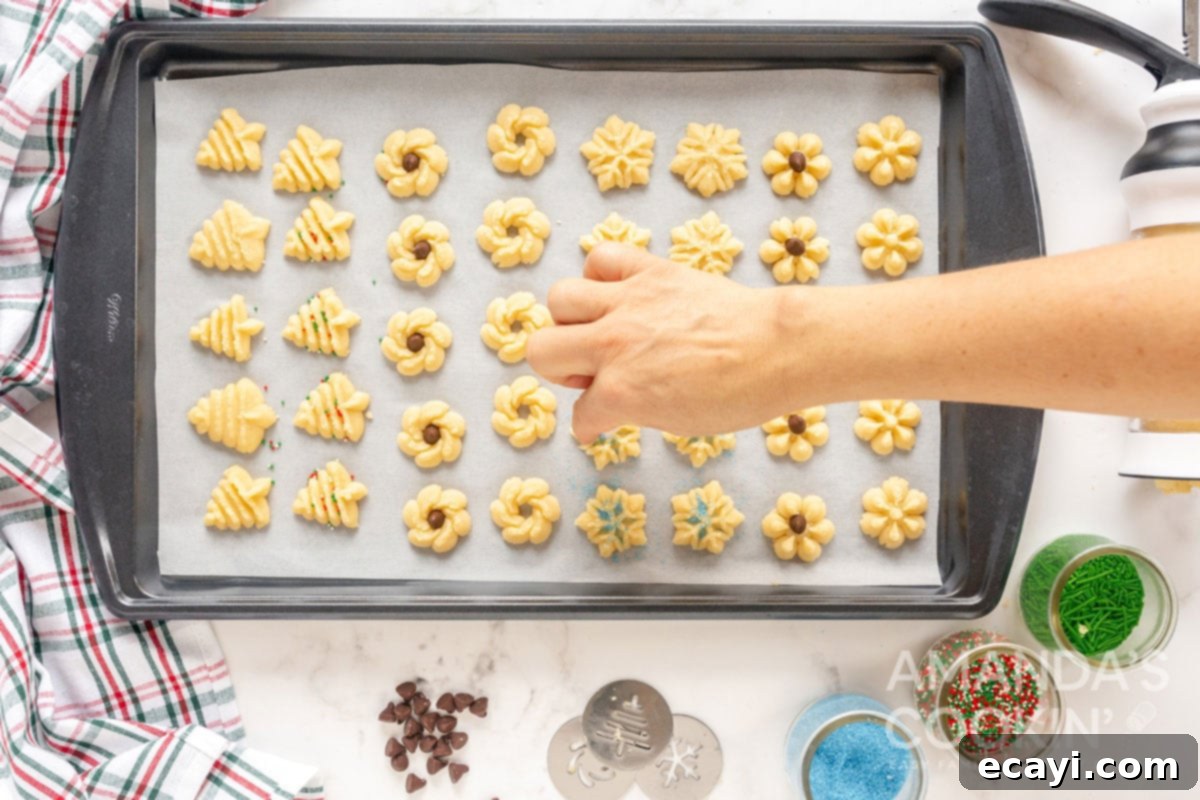 Spritz Cookies 10 A hand gently sprinkling vibrant colored sugar over newly pressed Spritz cookies on a baking sheet, adding a festive touch.