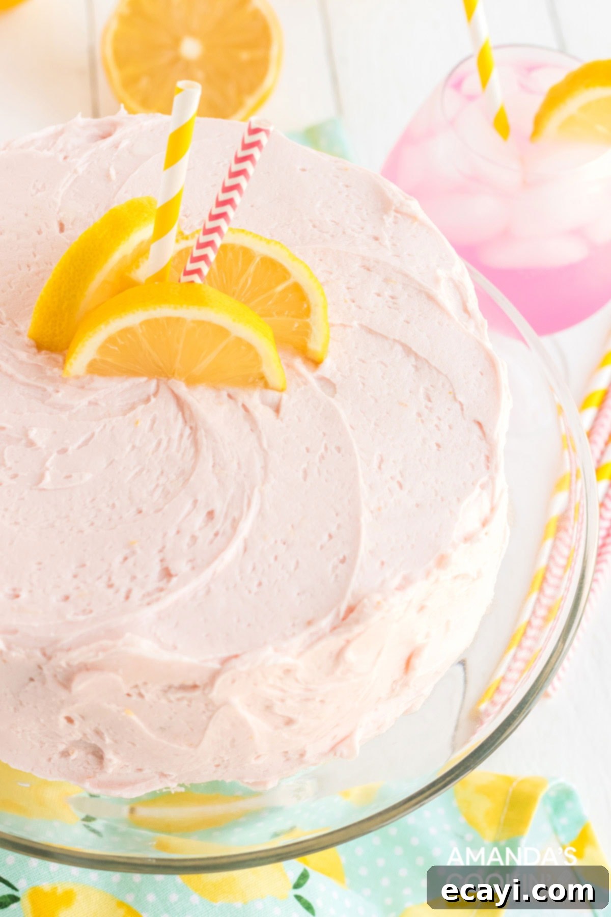 This pink lemonade cake is perfect for summer and will be a huge hit with lemonade fans. Make this beautiful cake from scratch for your next party! A beautifully frosted two-layer pink lemonade cake, garnished with lemon slices, on a white platter ready to be served. Ideal for summer gatherings.