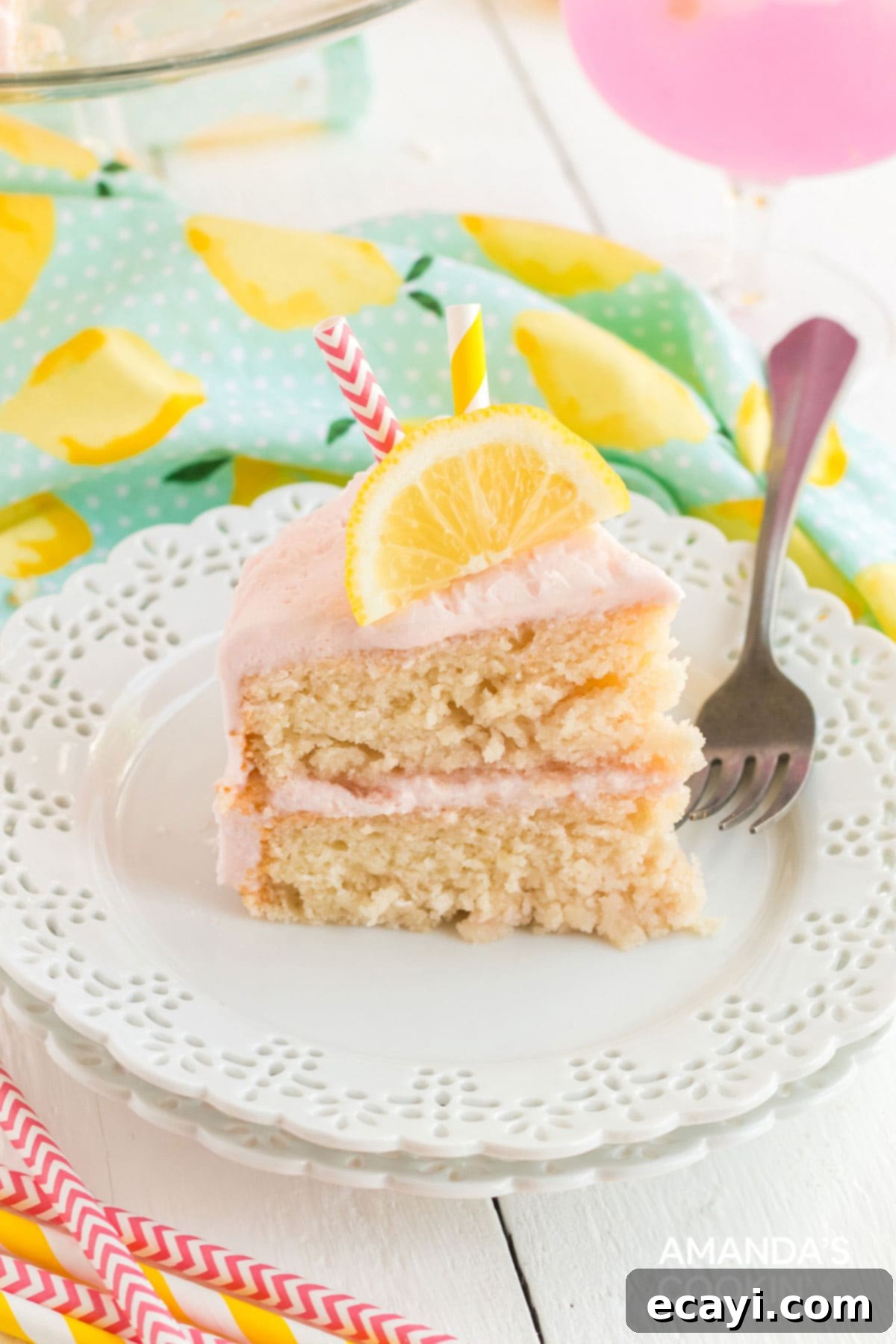 This pink lemonade cake is perfect for summer and will be a huge hit with lemonade fans. Make this beautiful cake from scratch for your next party! A perfect slice of homemade pink lemonade cake on a white plate, showcasing its fluffy layers, pink frosting, and a fresh lemon slice garnish.