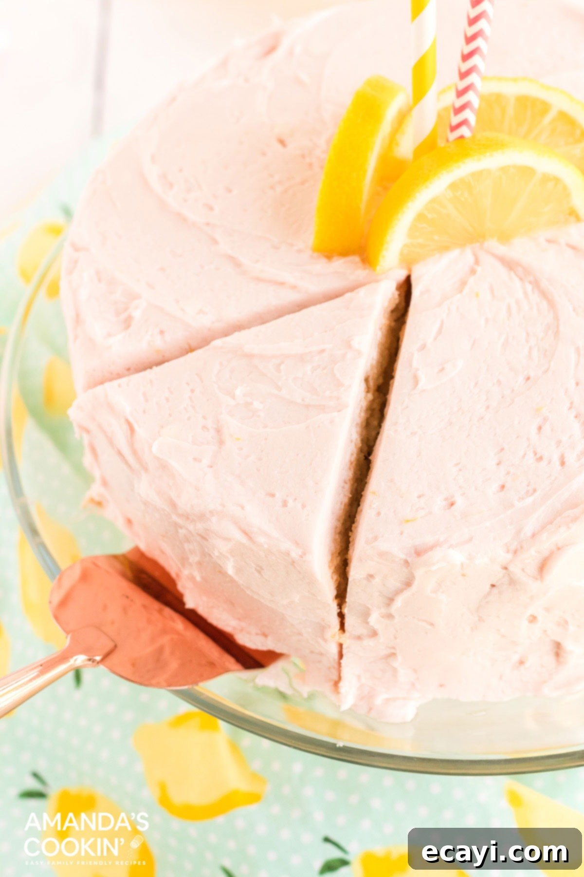 This pink lemonade cake is perfect for summer and will be a huge hit with lemonade fans. Make this beautiful cake from scratch for your next party! A hand using a pie server to remove a slice of moist, round pink lemonade cake from a serving platter, showing its fluffy texture and pink frosting.