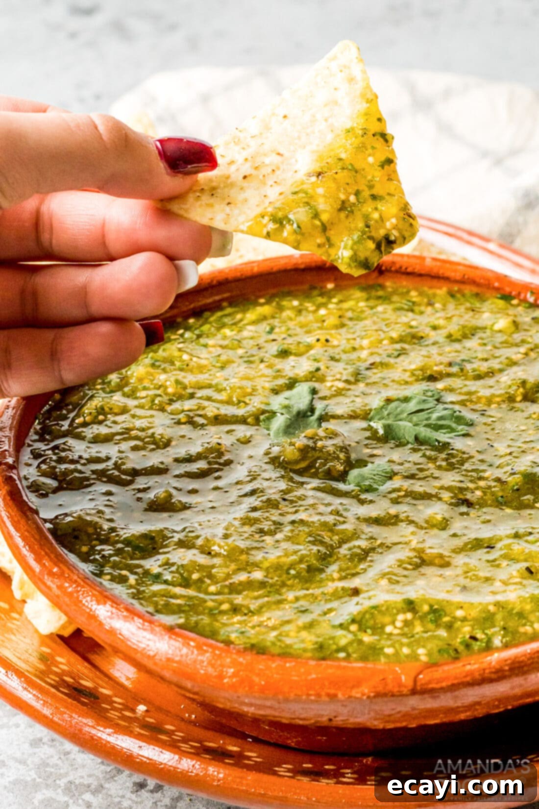Green Salsa 3 A woman's hand gracefully scoops up a generous portion of vibrant green salsa verde with a golden tortilla chip from a white bowl, showcasing its perfect consistency.