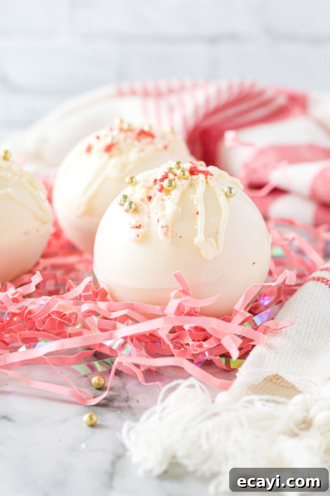 Dreamy Strawberry White Chocolate Hot Cocoa Bombs 2 Strawberry White Chocolate Hot Cocoa Bombs beautifully arranged on pink shredded paper, showcasing their delicate pink hue and inviting appearance. Perfect for Valentine's Day gifts or party favors.