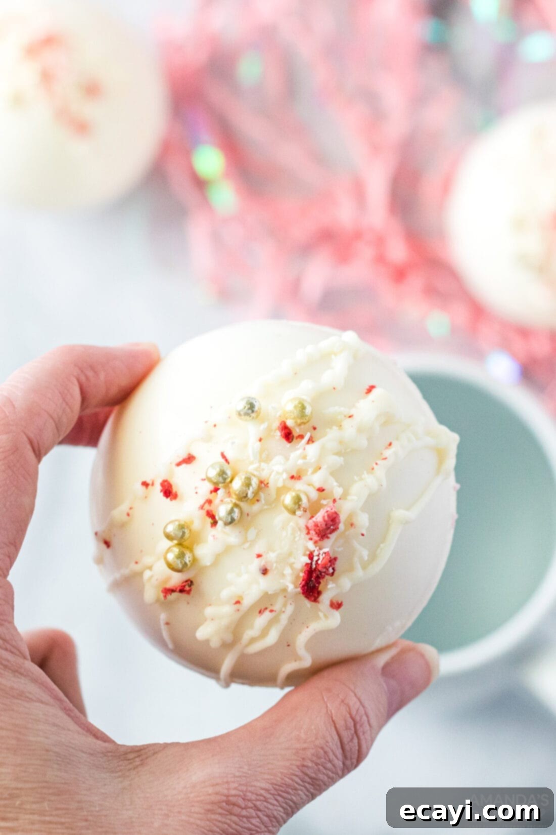 Dreamy Strawberry White Chocolate Hot Cocoa Bombs 3 A hand carefully holds a strawberry white chocolate hot cocoa bomb over a mug, ready to be dropped into hot milk or water for a delicious drink.
