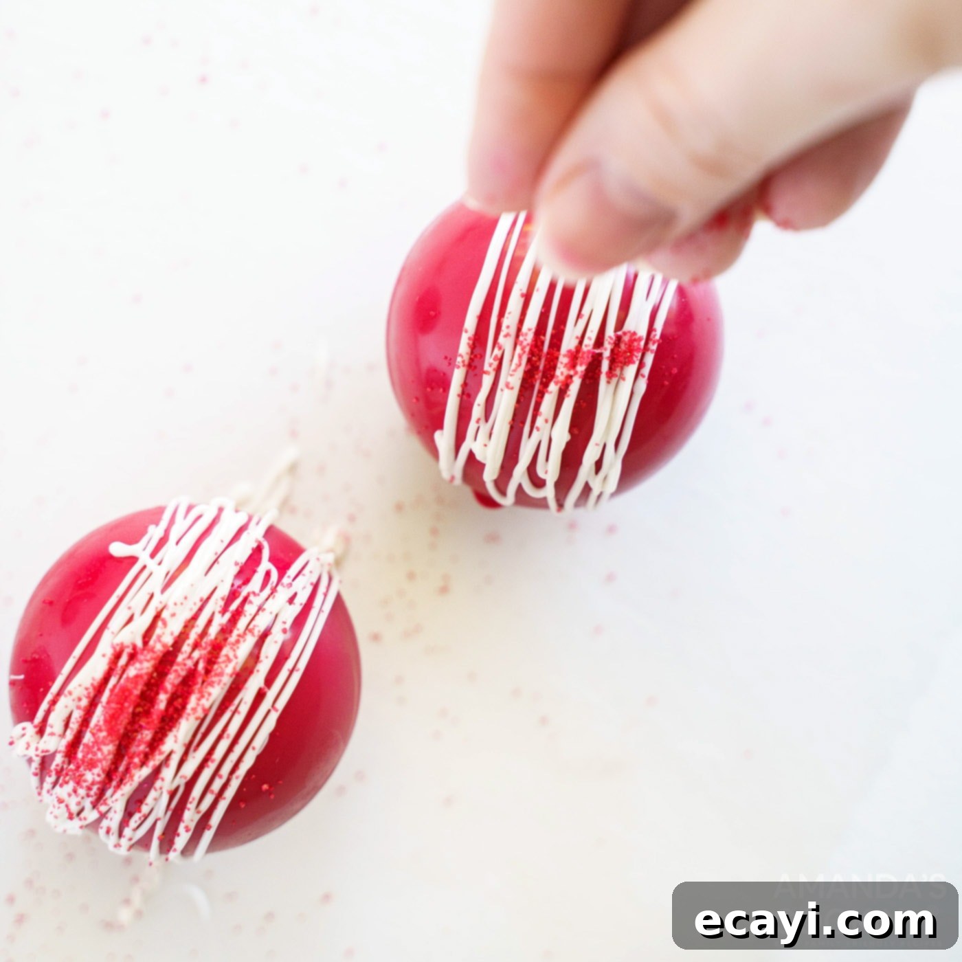 Decadent Red Velvet Hot Cocoa Bombs 13 A close-up shot of a hand adding colorful sprinkles onto freshly drizzled white candy on red velvet cocoa bombs.