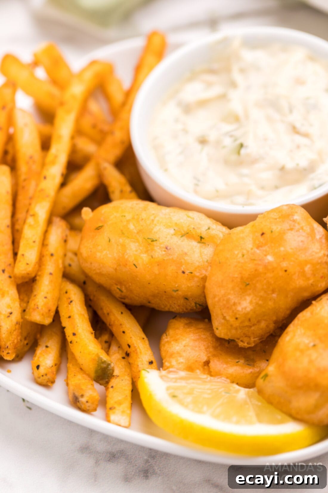Crispy Golden Beer-Battered Fish 2 Crispy beer battered fish and golden french fries served with a side of homemade tartar sauce and a lemon wedge on a white plate, ready to be enjoyed.