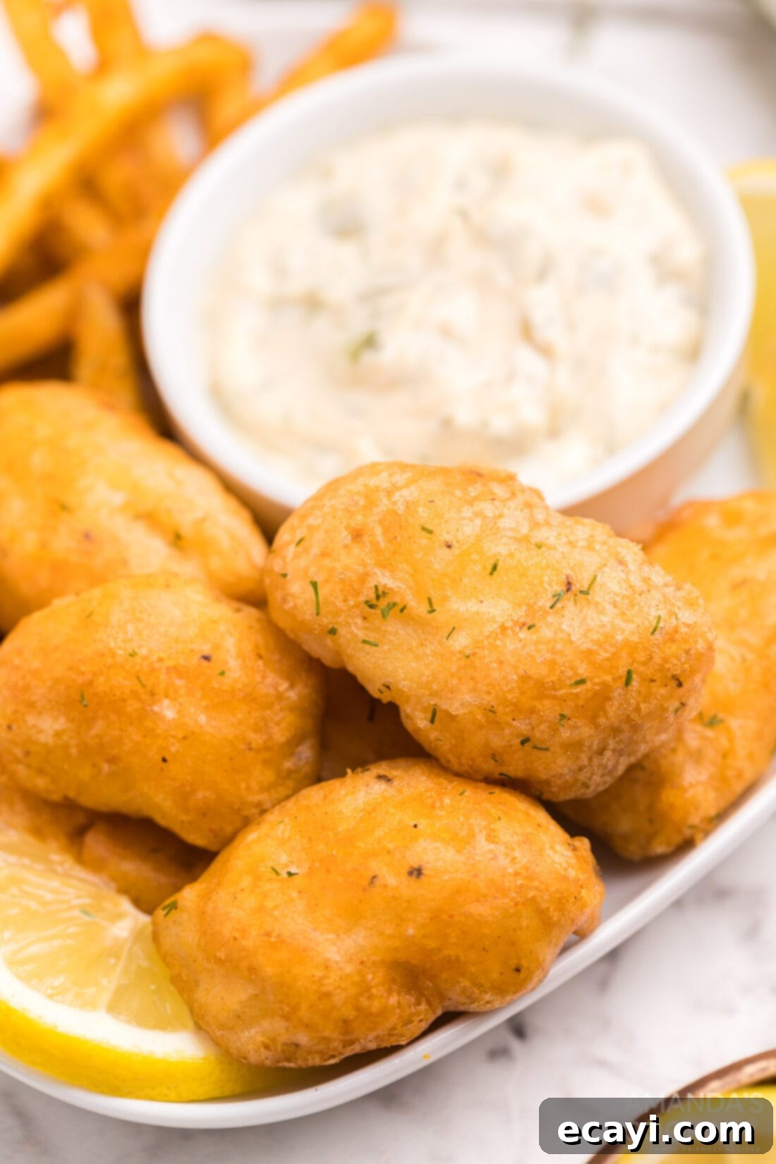 Crispy Golden Beer-Battered Fish 10 A plate featuring perfectly golden and crispy beer battered fish nuggets, alongside a small bowl of creamy homemade tartar sauce, a lemon wedge, and a sprinkle of fresh herbs.
