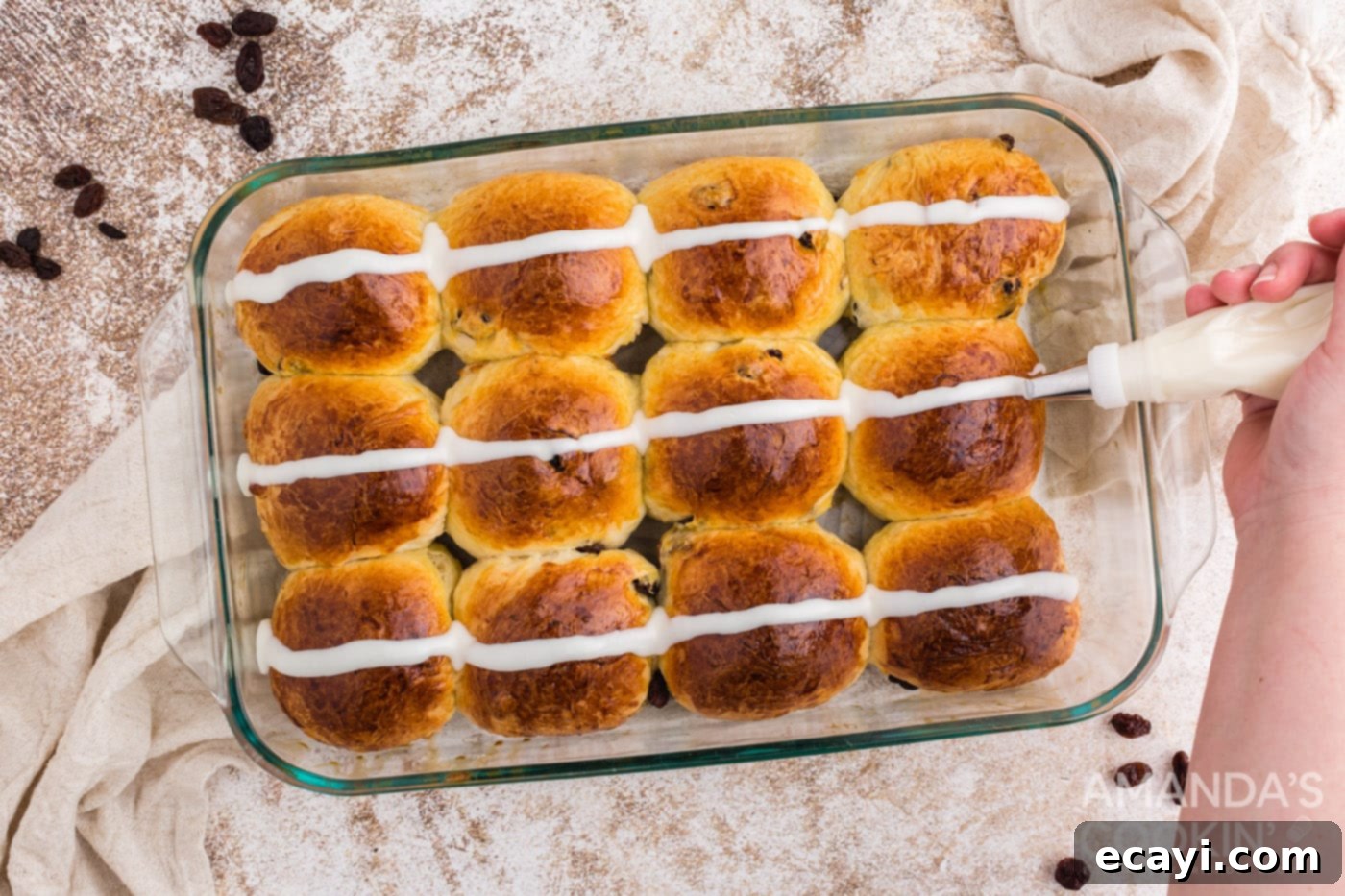 Spiced Easter Buns 17 A piping bag applying white icing in horizontal strips across baked Hot Cross Buns in a pan, forming the cross pattern.