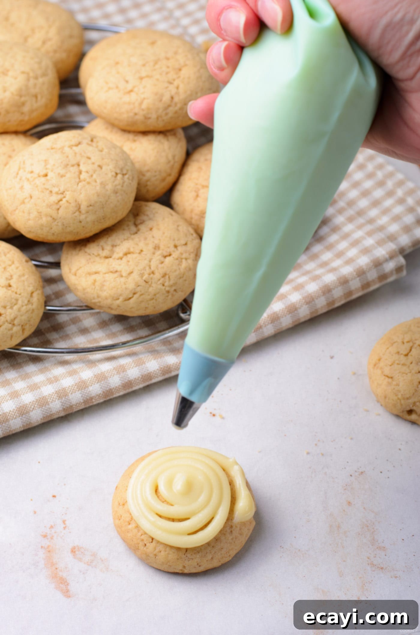 Cinnamon Swirl Delights 9 Piping a creamy, white cream cheese frosting in a spiral pattern onto a cooled cinnamon roll cookie, with a sprinkle of cinnamon on top.