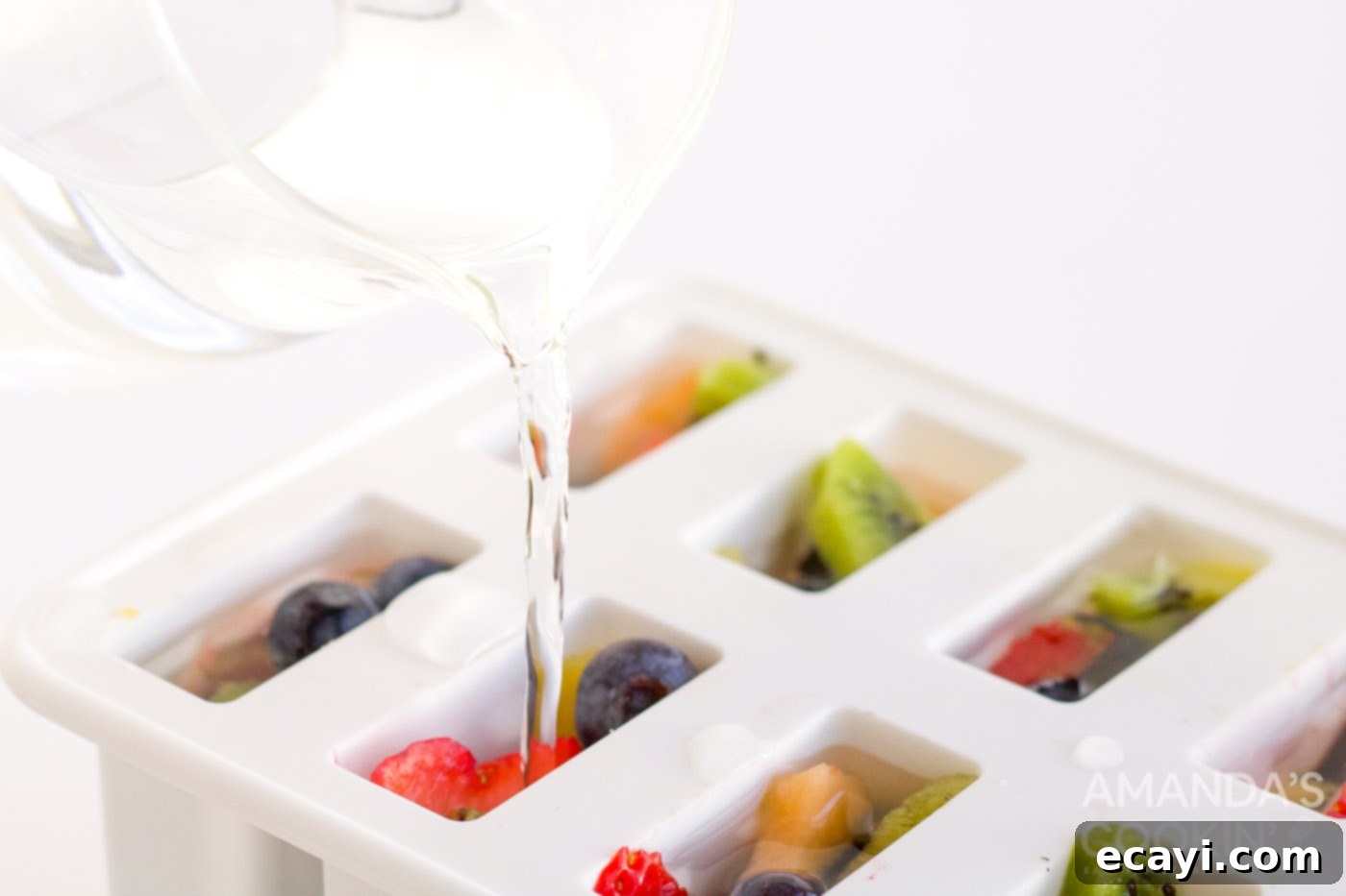 Refreshing Fruit Treats 6 A hand pouring white grape juice over colorful fresh fruit inside clear popsicle molds, preparing them for freezing.