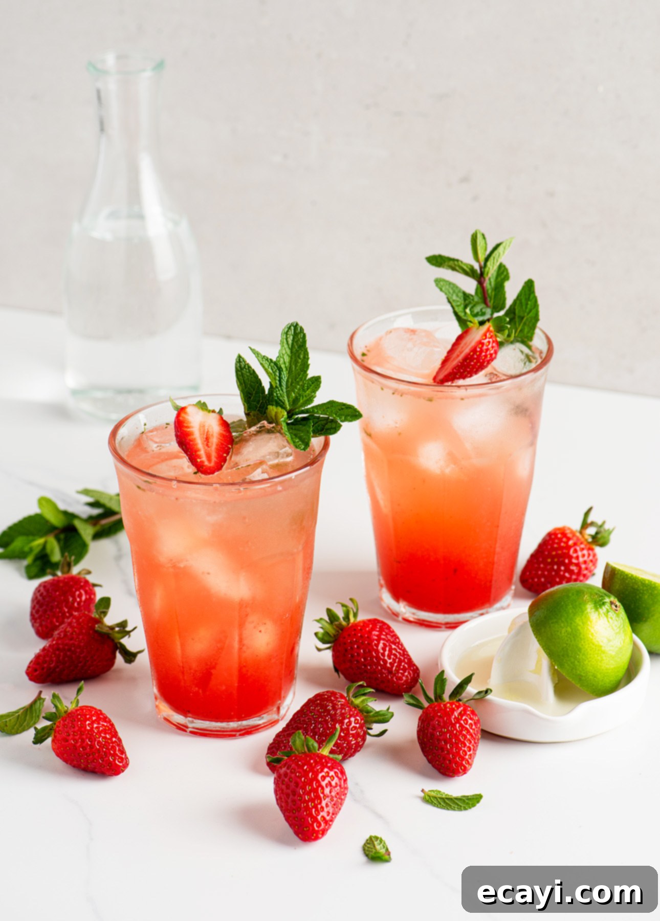 Strawberry Mojito Bliss 2 Two glasses of strawberry mojito cocktail with ice, garnished with fresh strawberries and mint, surrounded by whole strawberries, lime halves, and a small carafe of water on a white surface. The perfect summer drink.