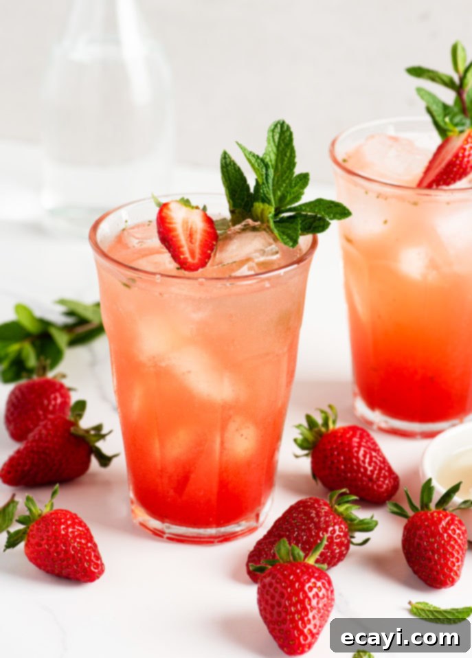 Strawberry Mojito Bliss 12 Two glasses of Strawberry Mojito Cocktail with ice, garnished with fresh mint and a sliced strawberry. Whole strawberries and mint leaves are scattered on a white surface around the beverages. The perfect refreshing summer drink.
