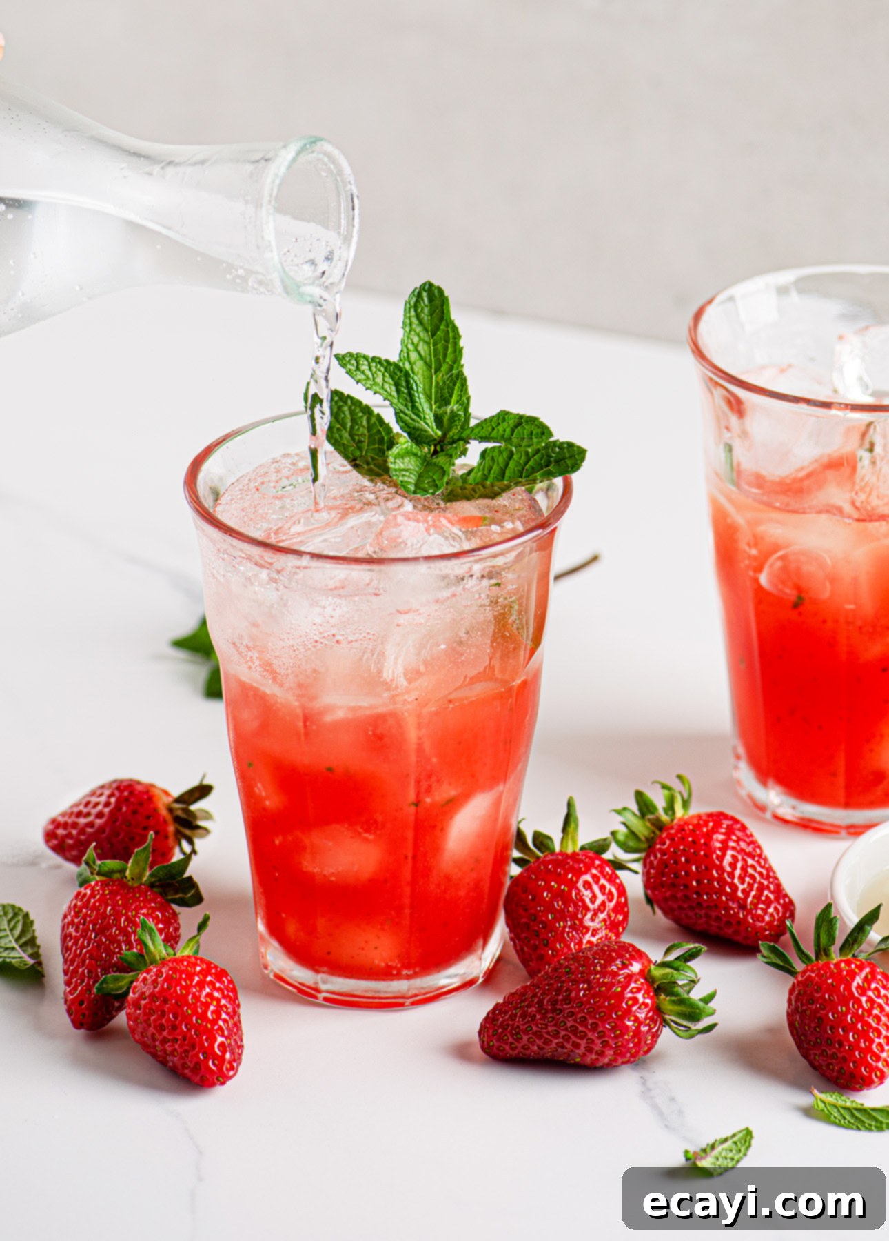 Strawberry Mojito Bliss 4 A glass of Strawberry Mojito Cocktail with ice, garnished with mint, is being topped with sparkling water. Fresh strawberries and mint leaves are scattered on a white surface, with another glass of the drink in the background. Capturing the essence of a refreshing summer drink.