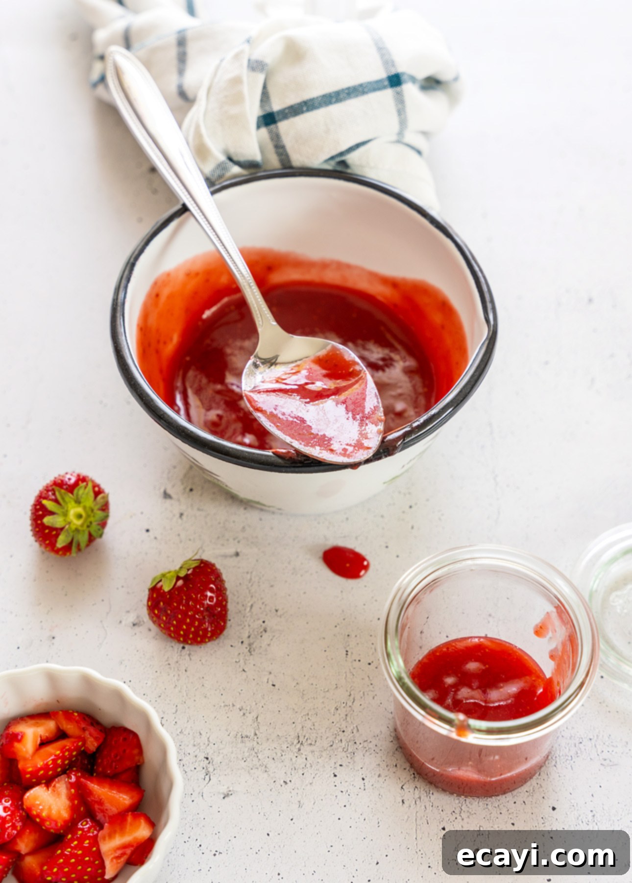 Strawberry Mojito Bliss 6 A bowl of strawberry puree with a spoon rests on a white surface. Nearby are fresh strawberries, a small dish of chopped strawberries, a jar of sauce, and a plaid kitchen towel—perfect for topping your next Strawberry Mojito or other desserts.
