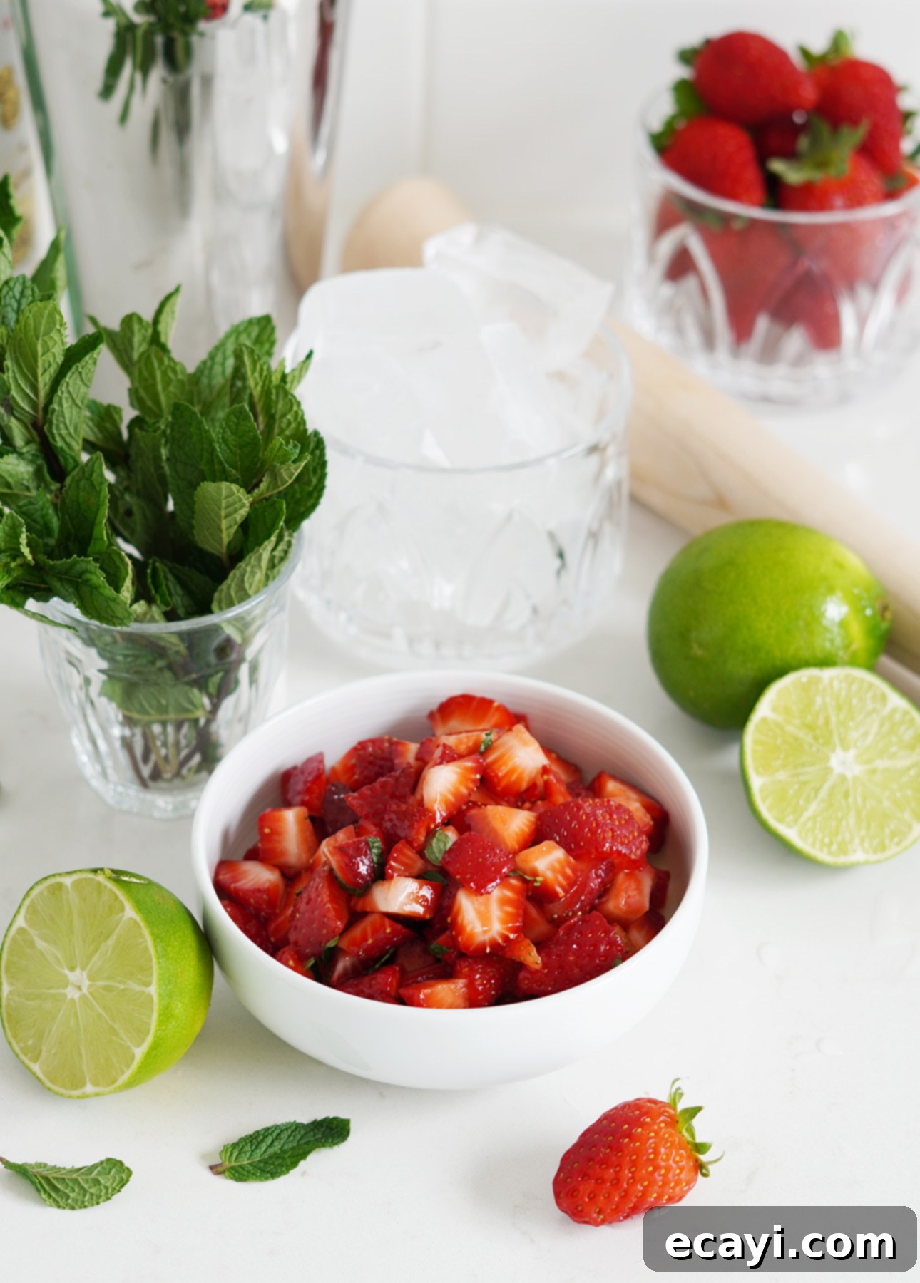Strawberry Mojito Bliss 9 A bowl of chopped strawberries with mint sits on a white surface, surrounded by fresh mint leaves, whole and halved limes, a strawberry, a glass of ice, and a container of whole strawberries—perfect for making a refreshing Strawberry Mojito Cocktail. Ingredients for a perfect summer drink.