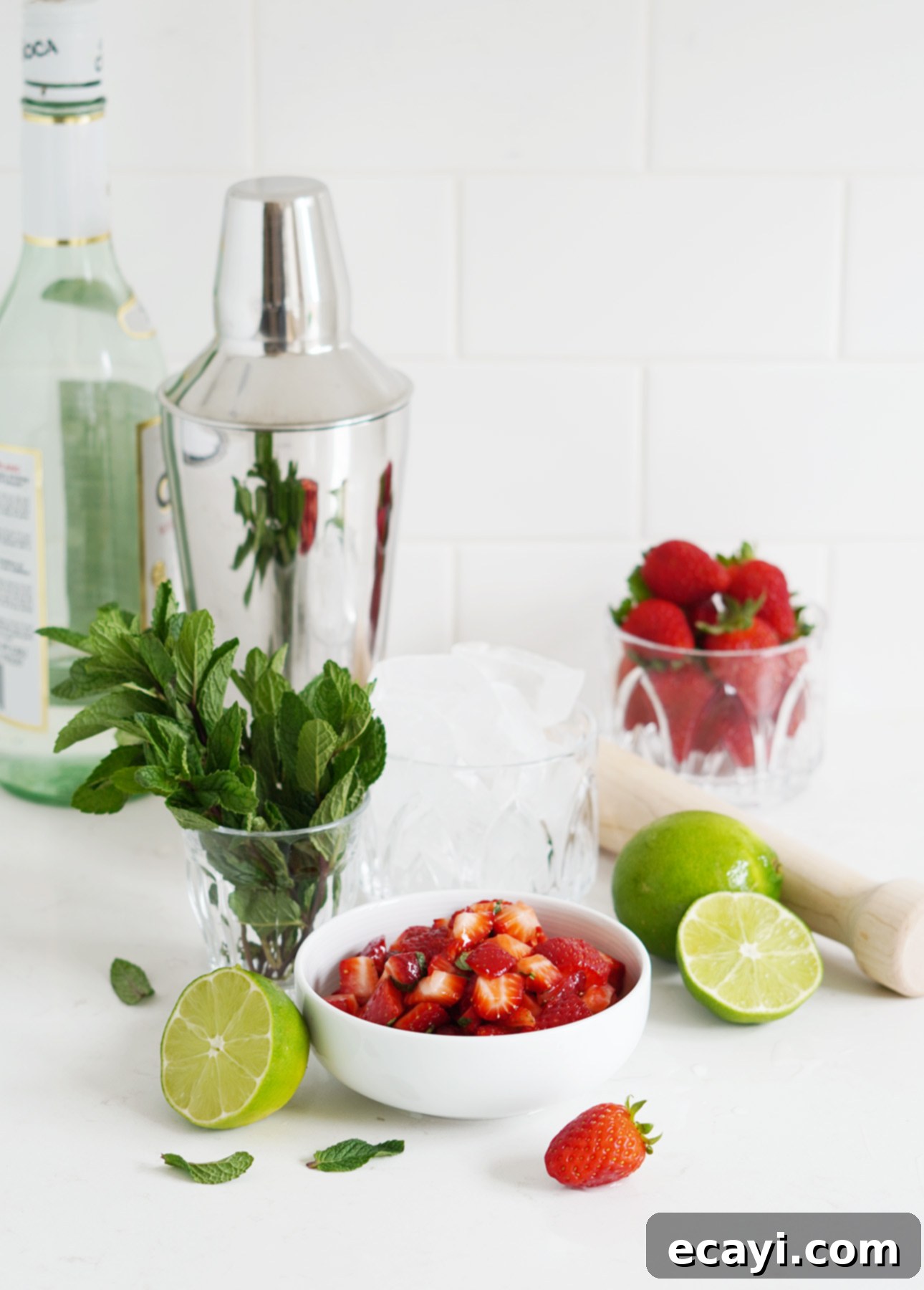 Strawberry Mojito Bliss 10 A white countertop with fresh mint, lime halves, chopped strawberries, a cocktail shaker, a glass of ice, a wooden muddler, a bowl of whole strawberries, and a bottle of clear alcohol—perfect for crafting a refreshing Strawberry Mojito Cocktail. All the necessary tools and ingredients.