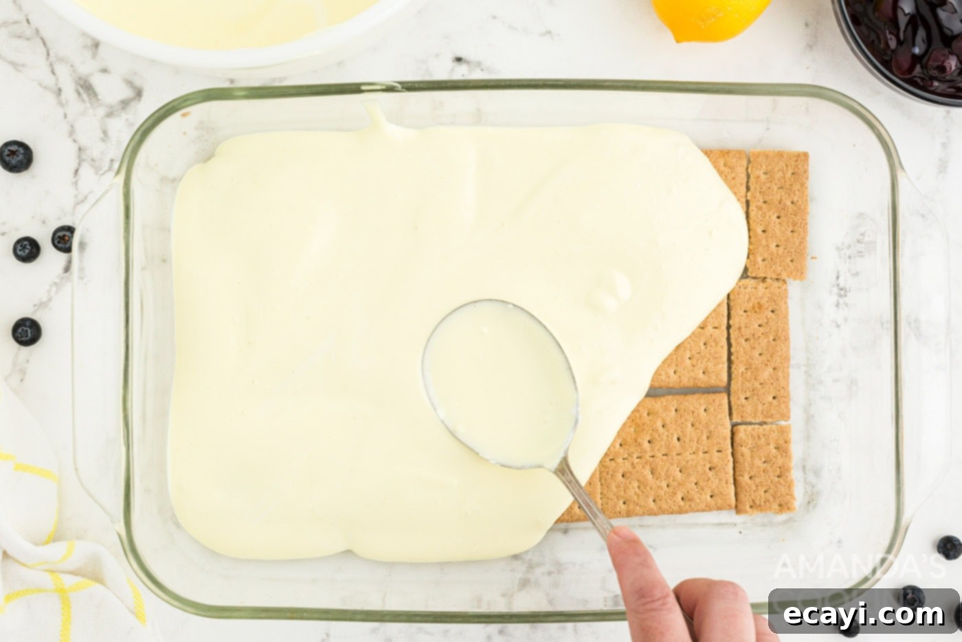 Zesty Lemon Blueberry Cloud Cake 7 A spoon spreading a thin layer of lemon pudding over graham crackers in a casserole dish.