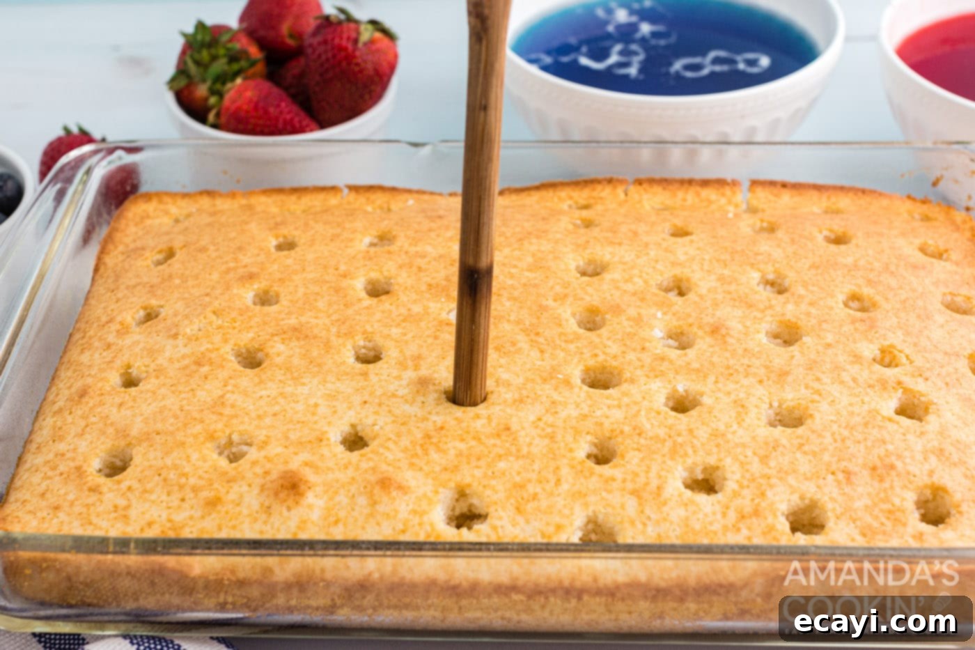 Patriotic Red, White and Blue Poke Cake 6 A hand using the handle of a wooden spoon to create even holes across a freshly baked white cake.