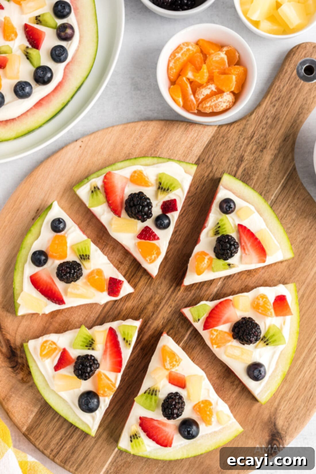 Watermelon Wonder Wheels 2 Watermelon Pizza on a cutting board, garnished with an assortment of fresh berries, kiwi, and mandarin oranges over a creamy yogurt spread.