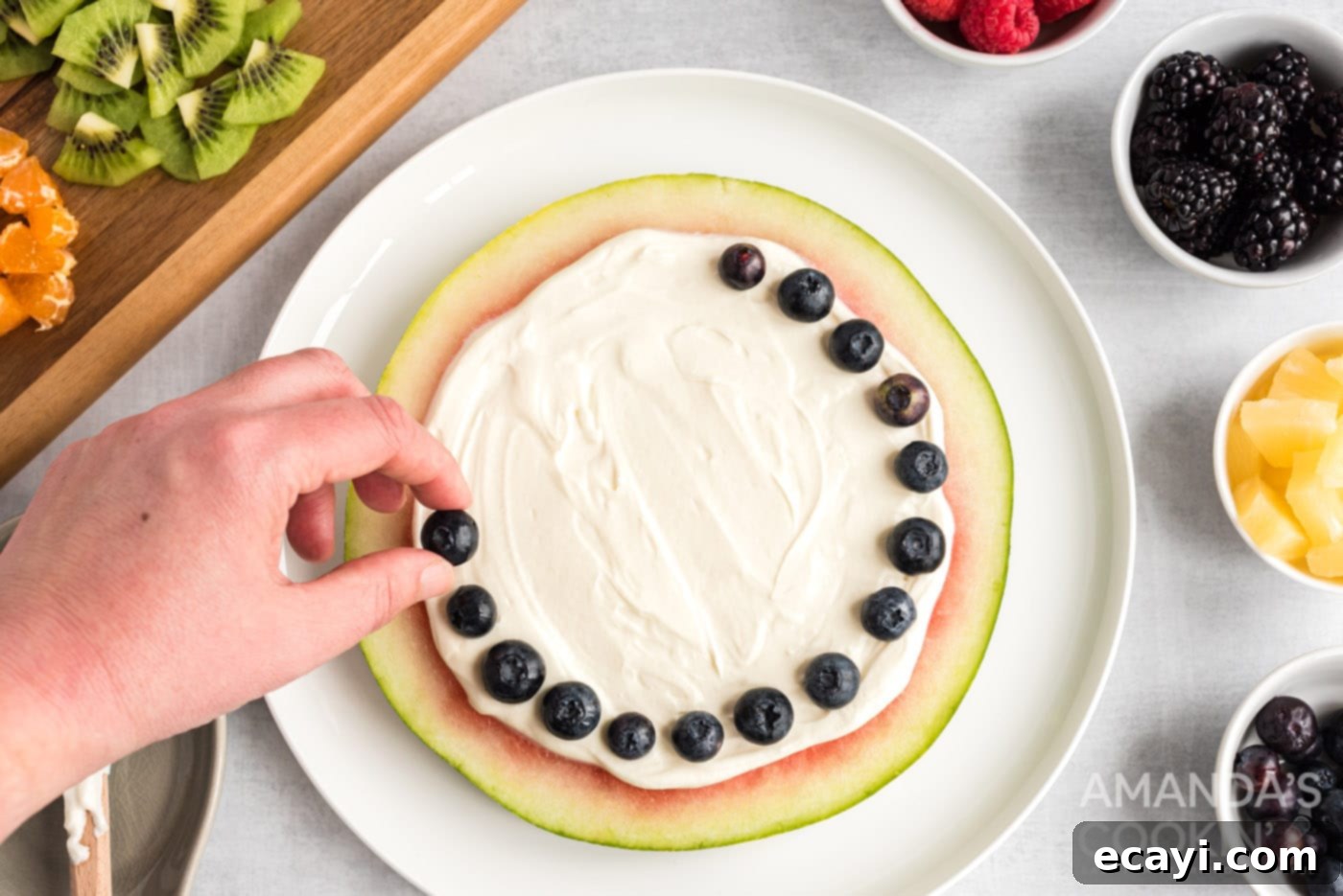 Watermelon Wonder Wheels 11 A hand carefully placing blueberries to form a perimeter around the cream cheese layer on a watermelon slice.