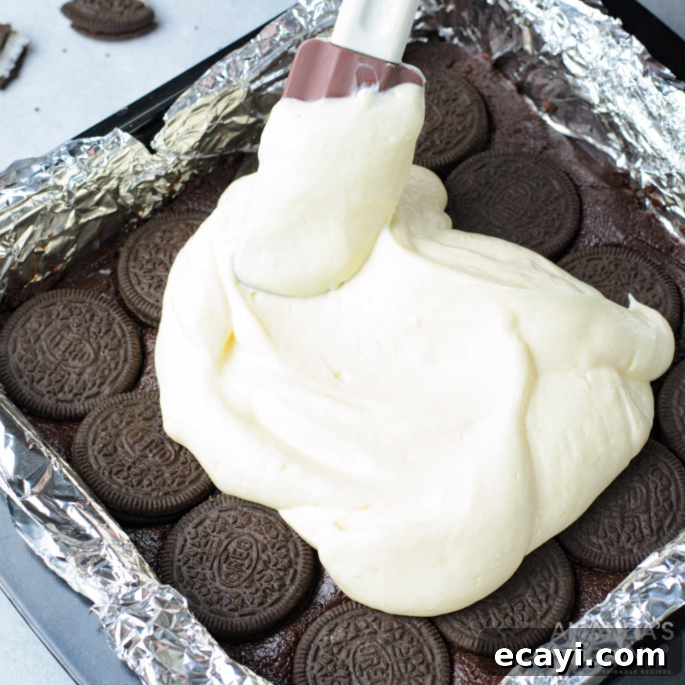 Decadent Oreo Cheesecake Brownies 11 A spatula smoothly spreading the creamy white cheesecake mixture over the layer of Oreo cookies and brownie batter in a baking pan.