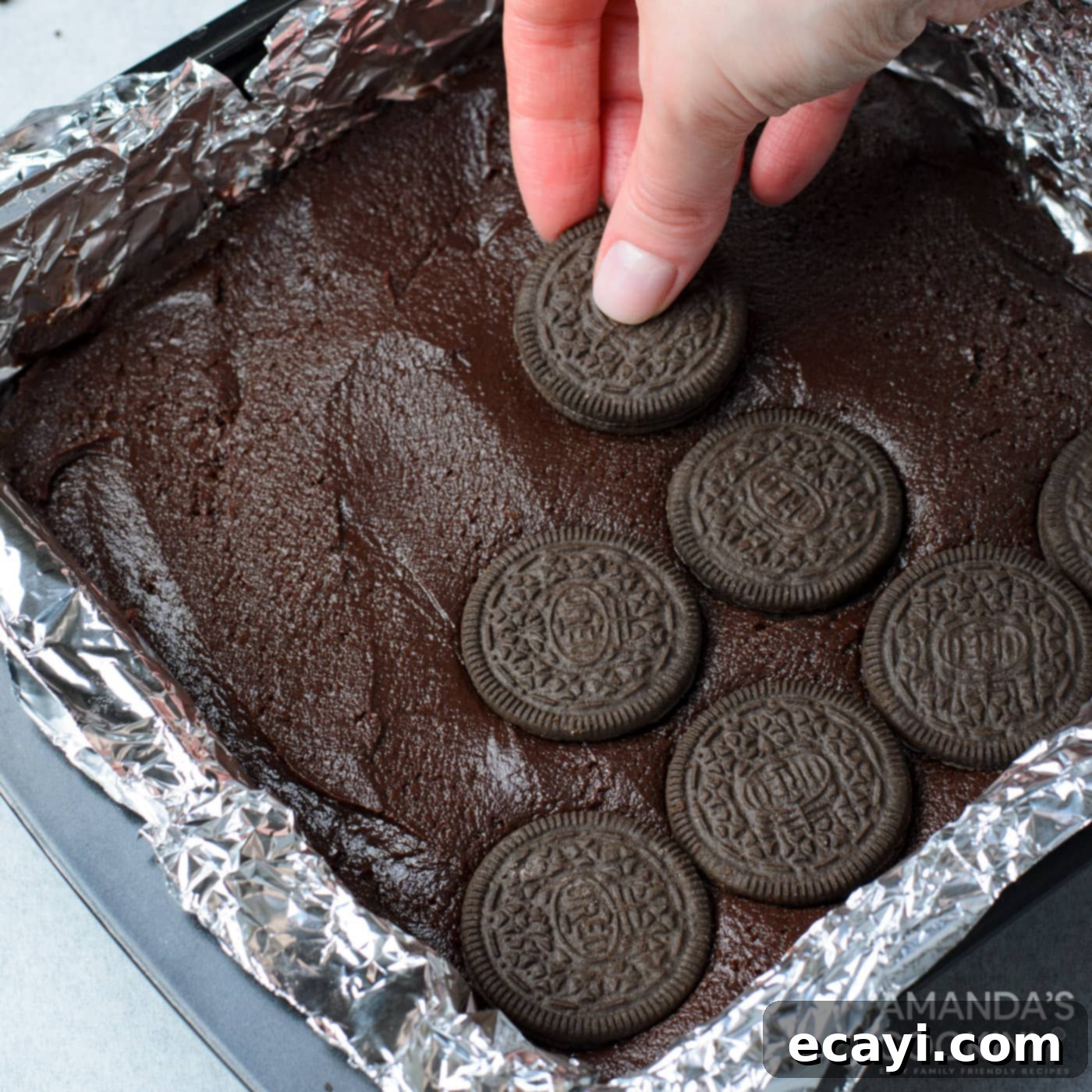 Decadent Oreo Cheesecake Brownies 10 A hand carefully placing whole Oreo cookies onto a smooth layer of brownie batter in a baking pan, preparing for the cheesecake topping.