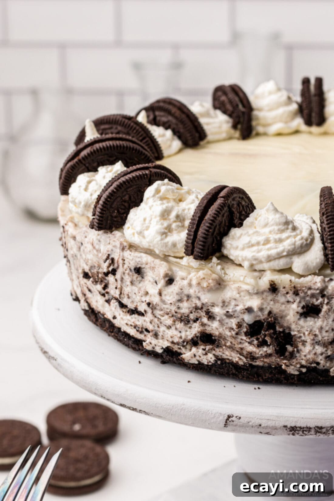 Decadent Oreo Cheesecake 2 A stunning whole Oreo Cheesecake on a white cake plate, featuring a creamy filling, cookie chunks, and a white chocolate ganache drizzle with whipped cream swirls and halved Oreos on top.