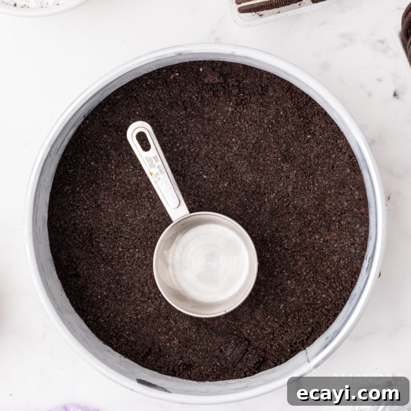 Decadent Oreo Cheesecake 6 A measuring cup being used to firmly press a layer of Oreo cookie crumbs into the bottom of a springform pan, creating a compact crust.