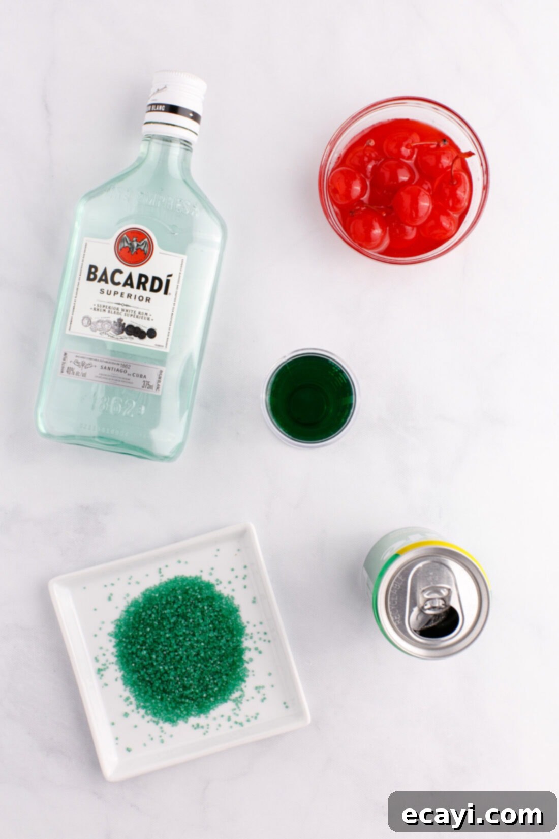 Grinch's Emerald Elixir 4 A flat lay showing all ingredients for making a Grinch Cocktail, including Midori, white rum, lemon-lime soda, cherries, and green sugar.