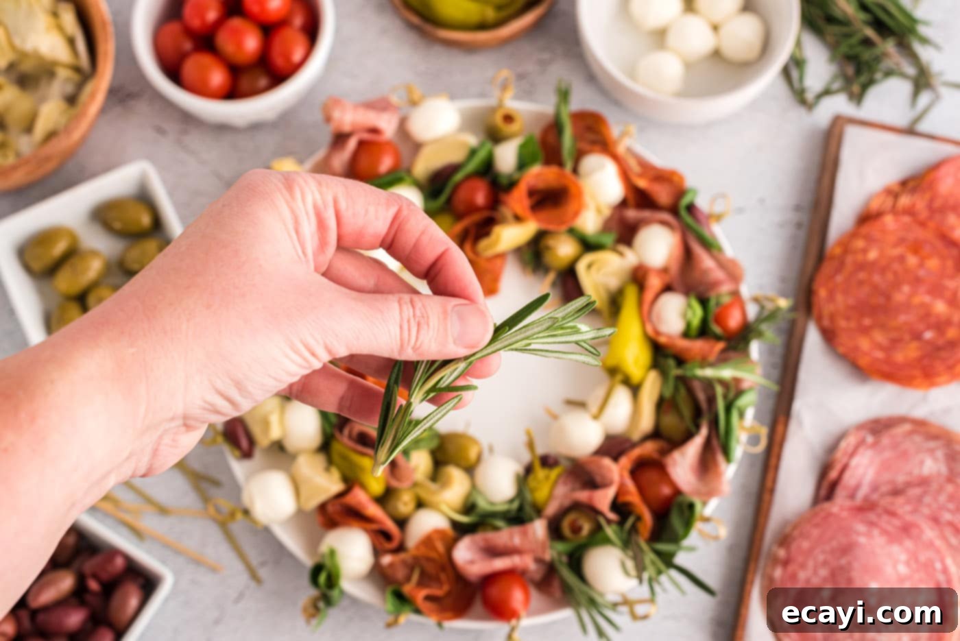 Gourmet Grazing Wreath 8 A hand placing a fresh rosemary sprig into the antipasto wreath, adding the finishing festive touch.