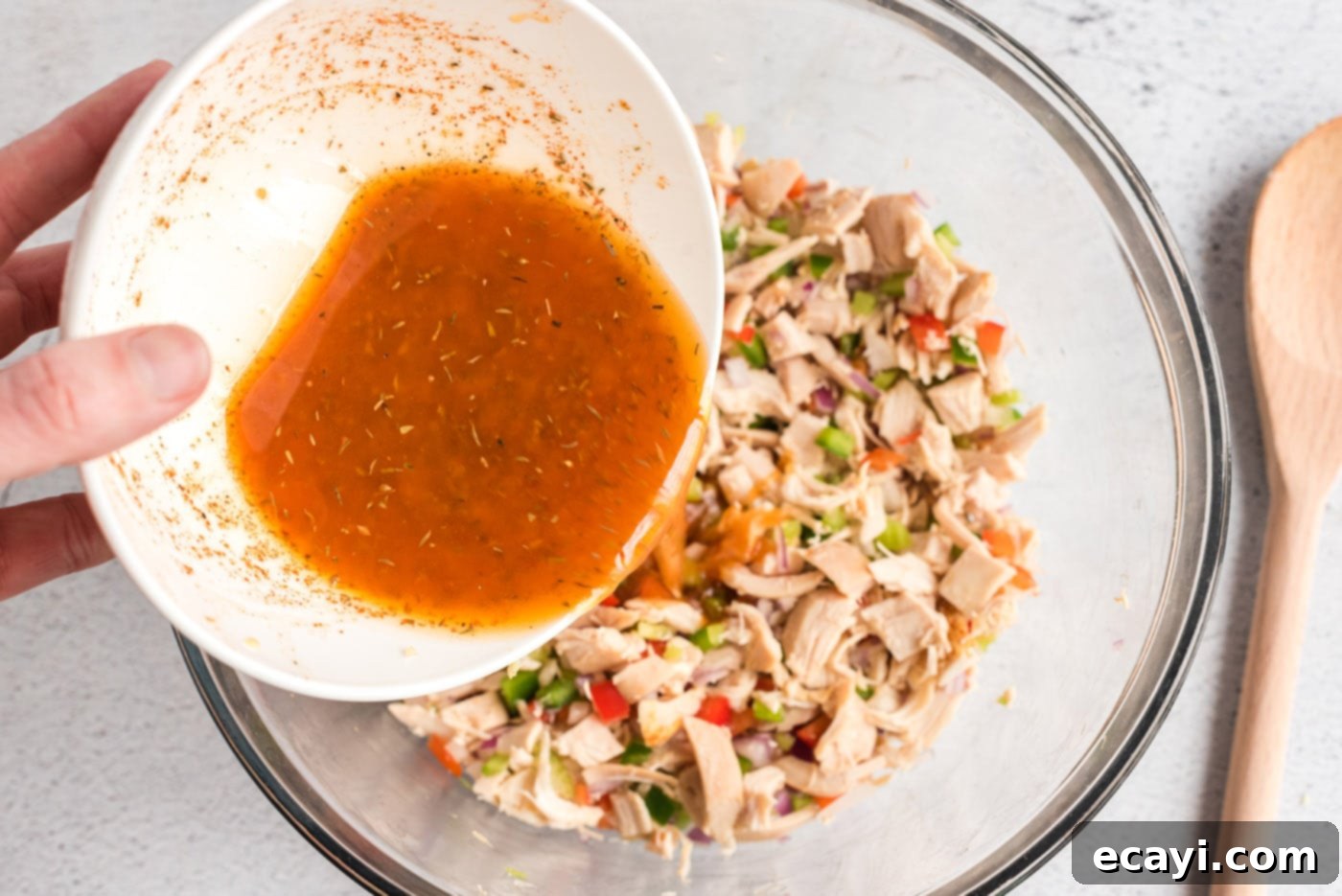 Bayou Chicken Salad 8 A stream of vibrant orange Cajun dressing being poured from a small bowl over the mixed chicken and vegetables in a large white mixing bowl, ensuring even coating.
