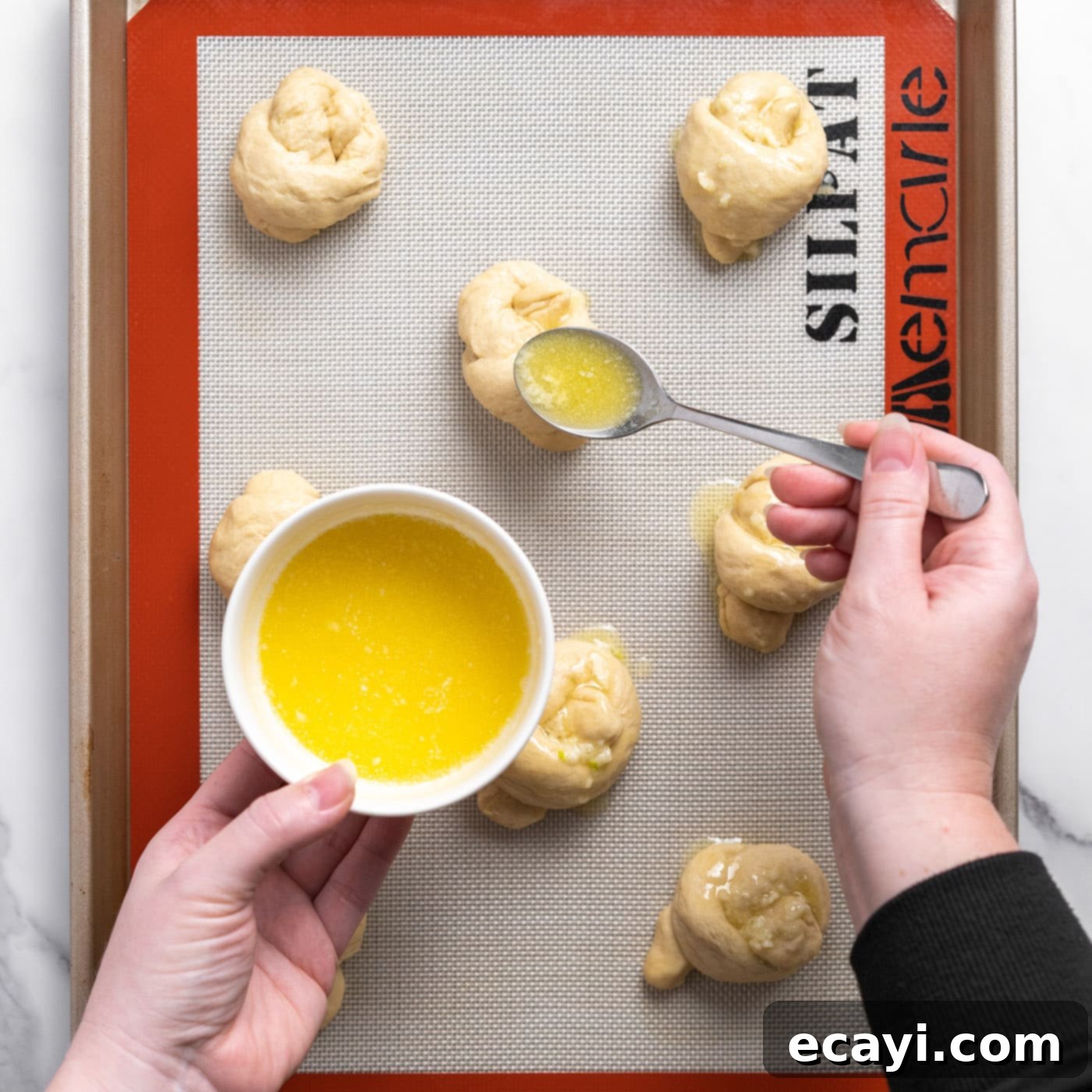 Pillowy Garlic Swirls 14 Melted garlic butter being spooned generously over unbaked dough knots arranged on a baking sheet, preparing them for the oven.