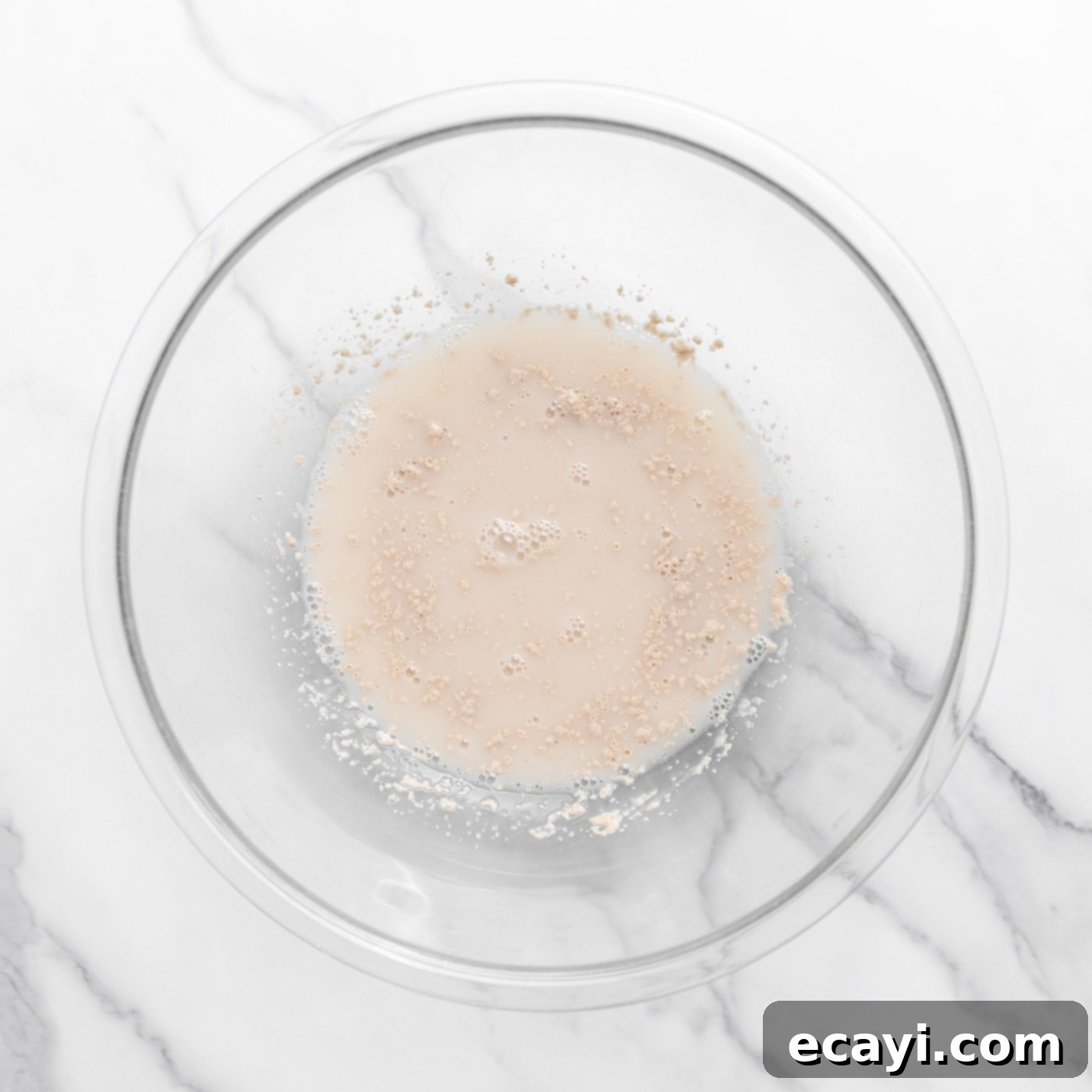 Pillowy Garlic Swirls 5 Warm water with instant yeast sprinkled on top, showing early signs of activation and foaminess in a glass bowl.