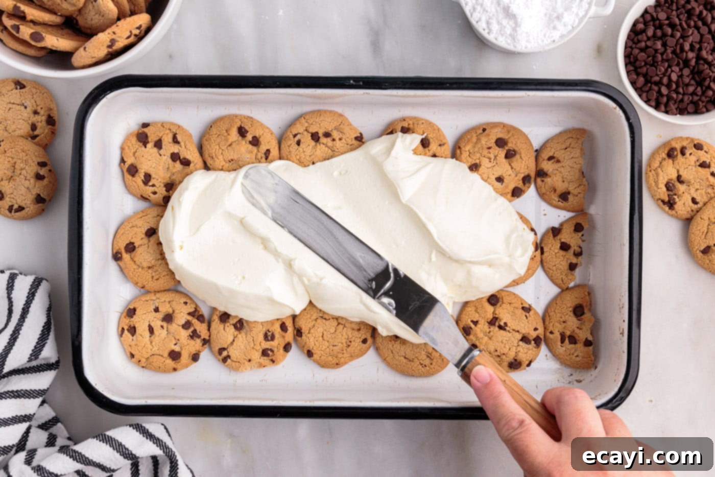 No-Bake Chocolate Chip Cookie Dream Cake 7 A layer of whipped cream cheese being spread smoothly on top of the chocolate chip cookies in a casserole dish.