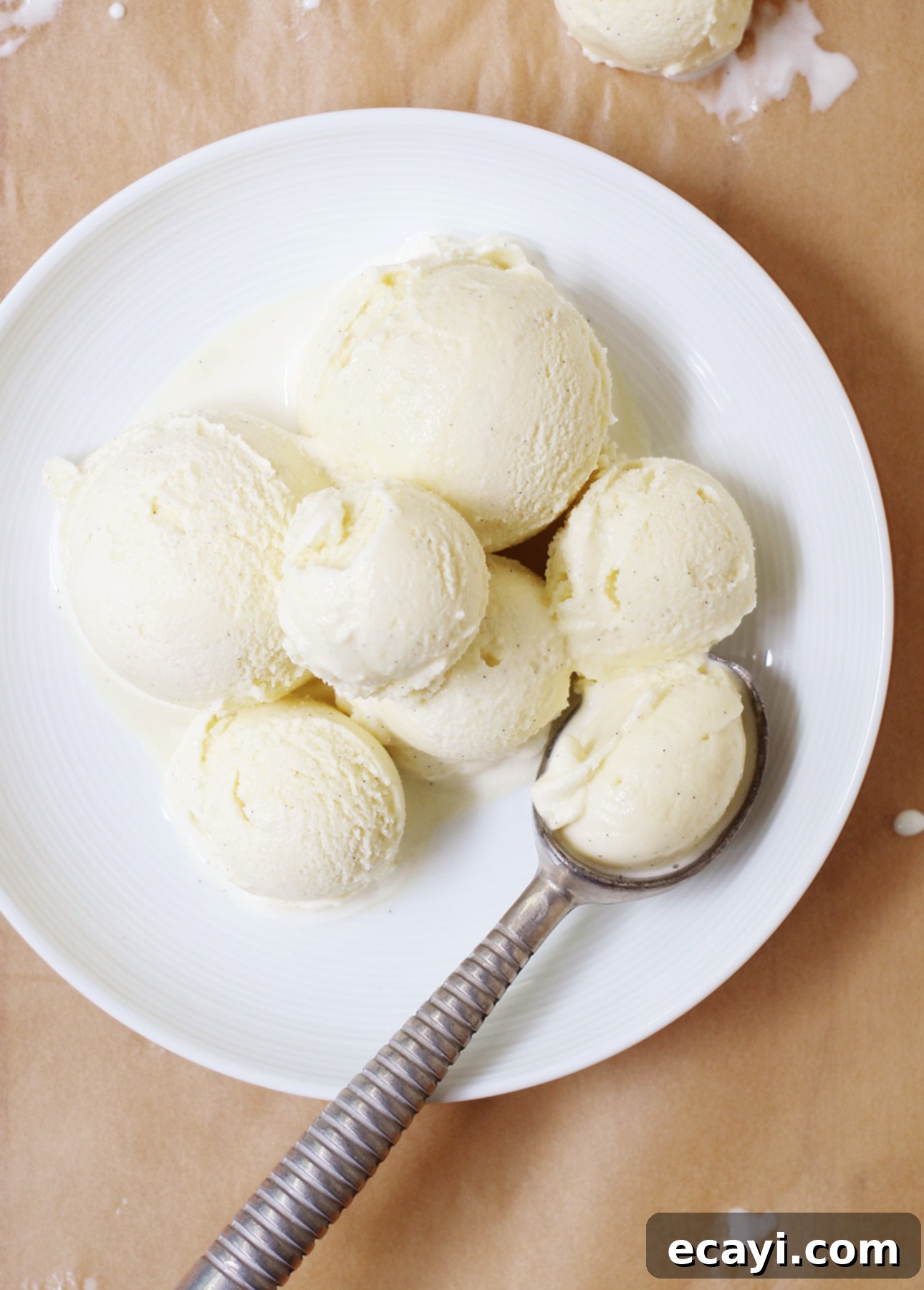 Homemade Italian Gelato: Simple Steps to Sweet Perfection 4 A white plate with several scoops of creamy vanilla gelato sits on a brown paper surface. A metal scoop, reminiscent of tools in How to Make Gelato guides, rests on the right side, cradling another scoop. Small drips dance around the plates edge. // FoodNouveau.com