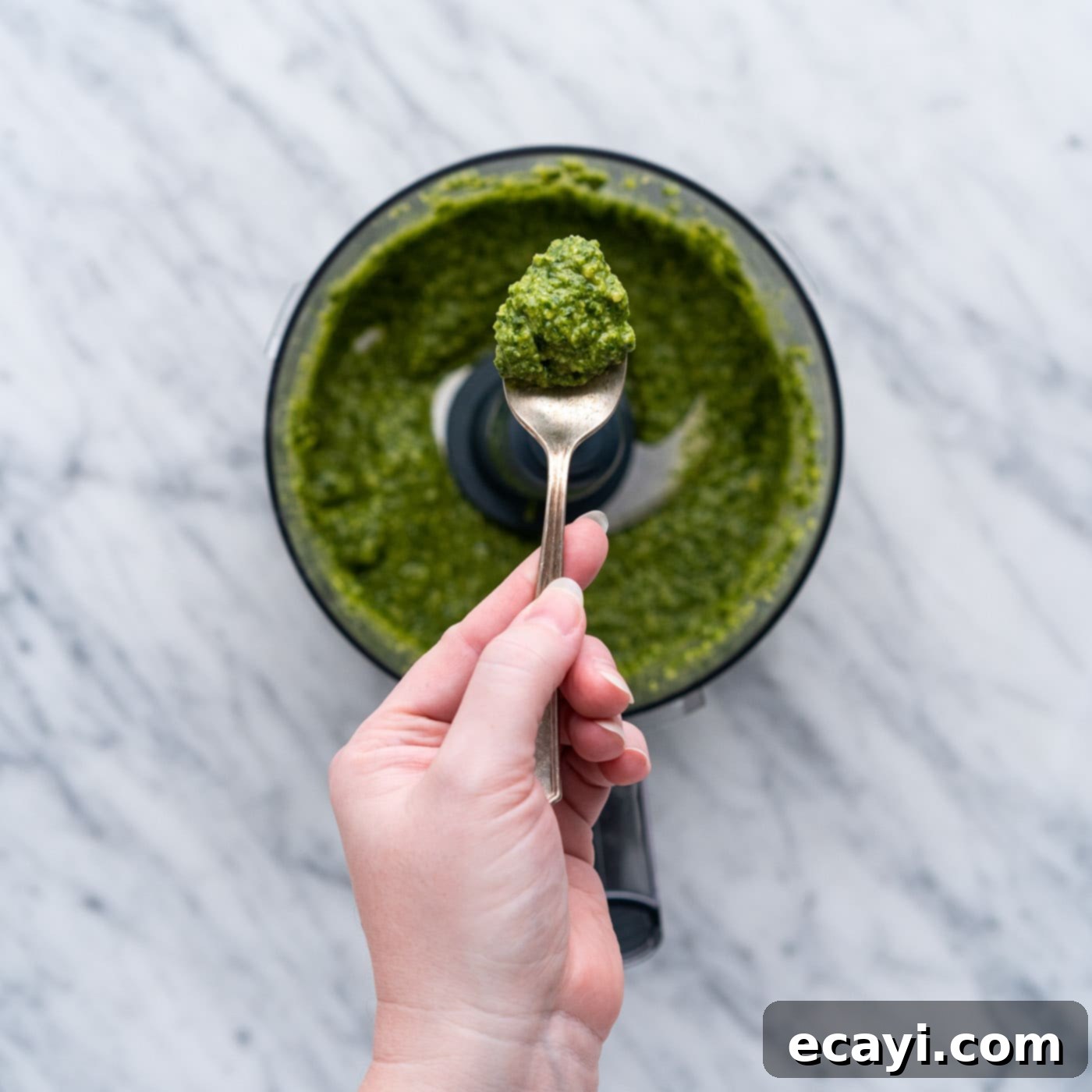 Pesto Perfection 7 A spoonful of freshly made basil pesto held above the food processor bowl, showing its rich green color and smooth texture.