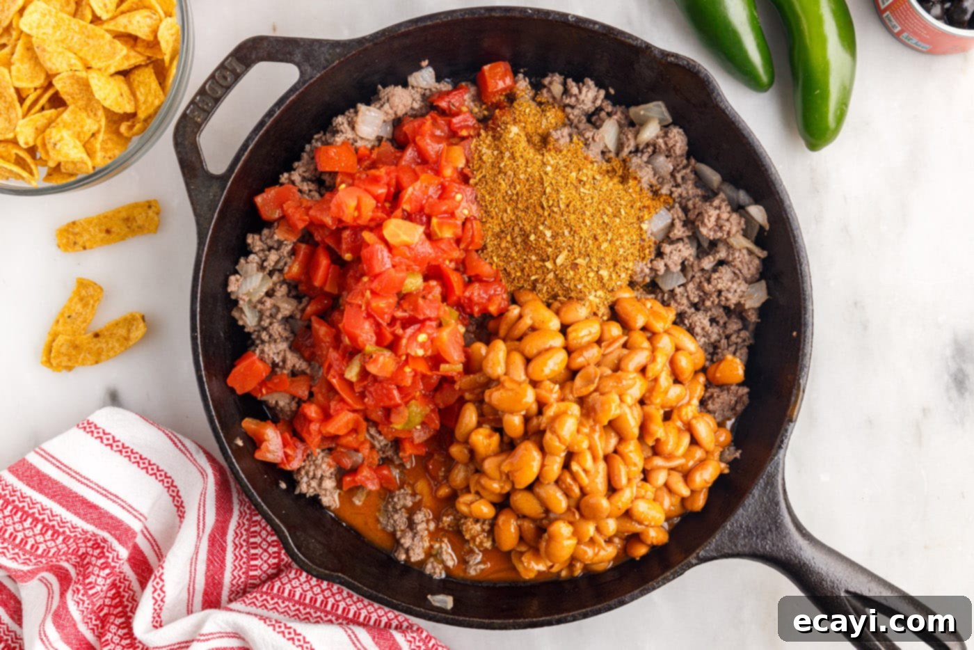 Crunchy Chili Cheese Delight 6 Taco seasoning, water, ranch-style beans, and Rotel tomatoes being stirred into the cooked ground beef and onion mixture in a skillet.