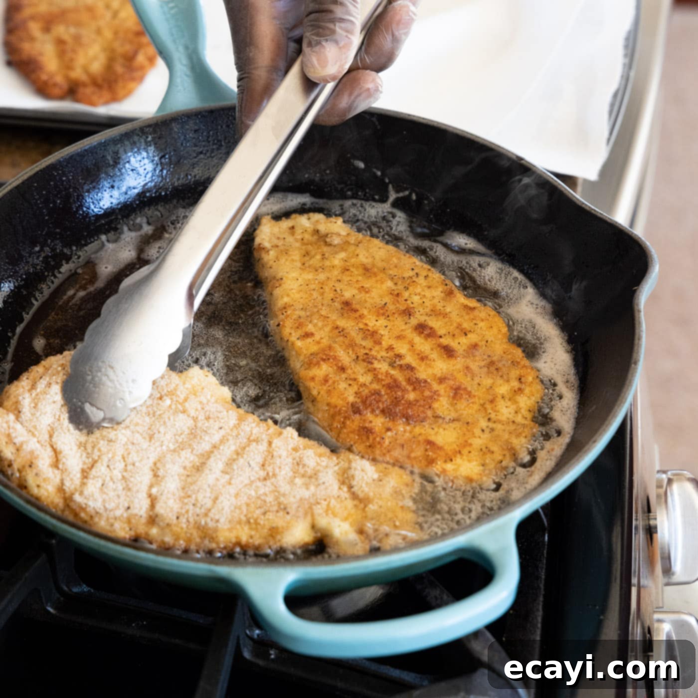 Crispy Chicken Milanese 13 Tongs flipping a chicken cutlet in a skillet, showing golden brown crust